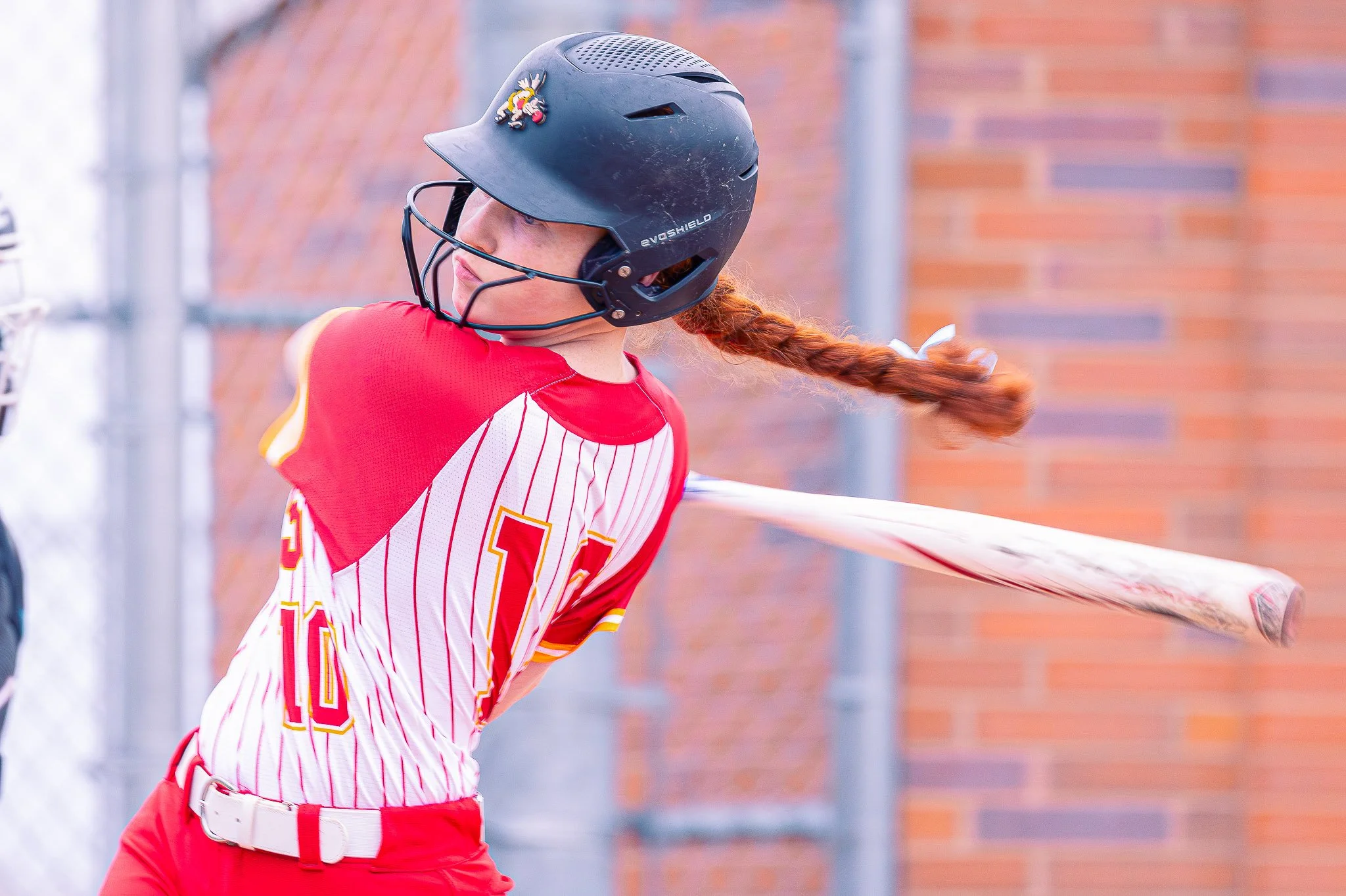 A female baseball player wearing a black batting helmet with a cartoon character pin, red and white striped jersey, and red pants, in the midst of batting during a game.