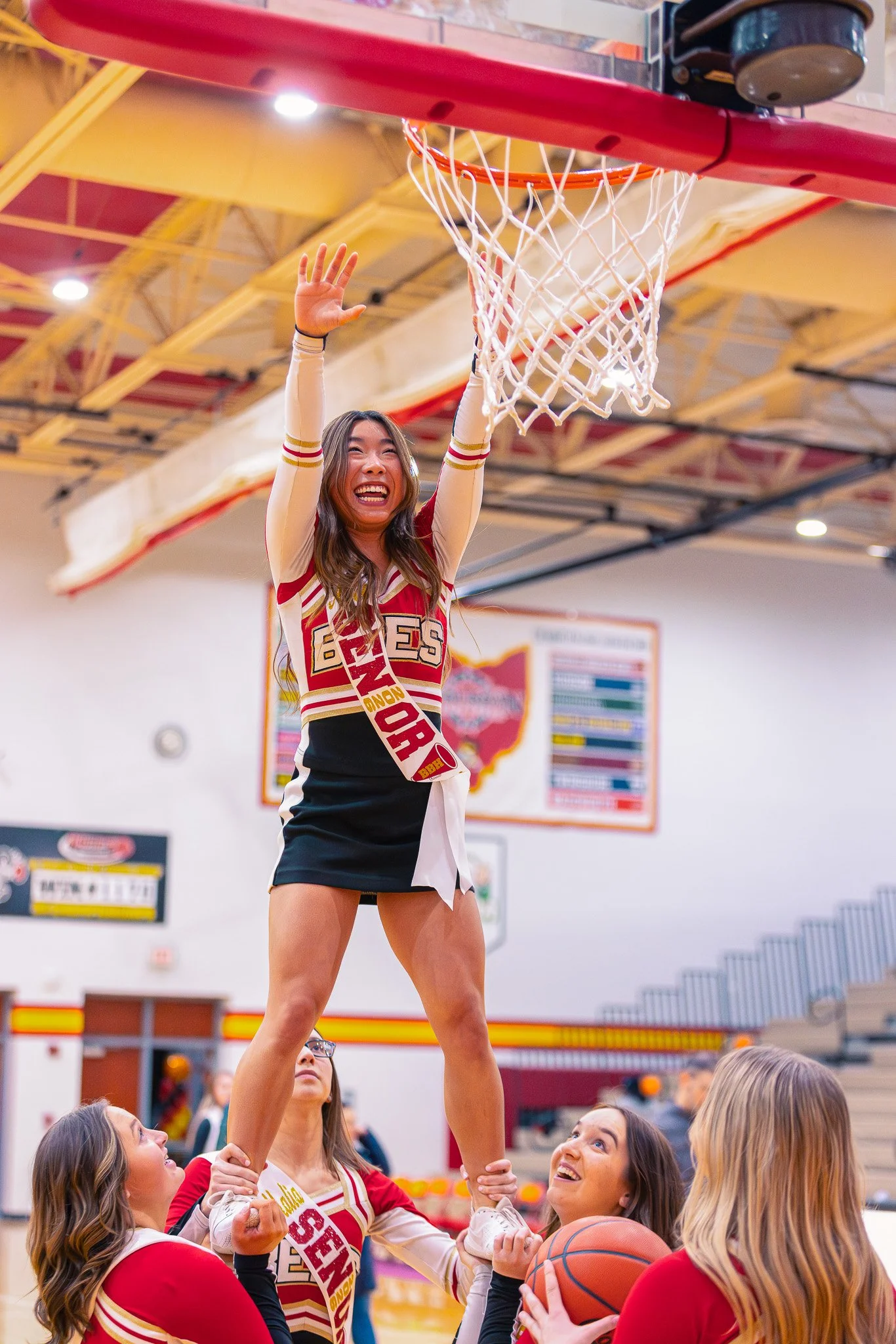 A cheerleader lifts a woman in a cheerleading stunt inside a gymnasium during a sports event. The cheerleader is smiling, wearing a cheerleading uniform and socks, and holding a cheerleading pose near the basketball hoop. Several other cheerleaders a