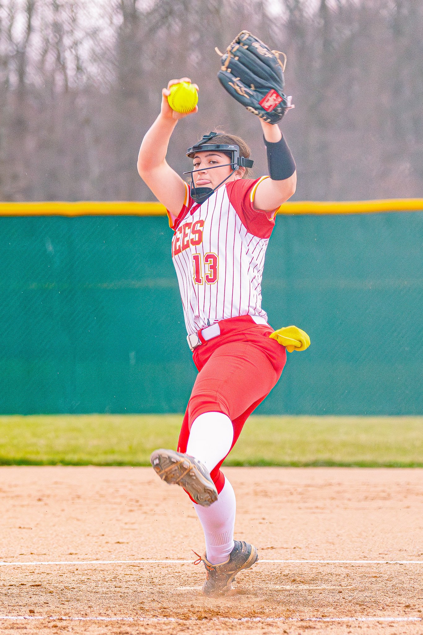 A female softball player wearing a red and white uniform with the number 13 is pitching on a softball field. She is in a dynamic pose, with one leg raised and arm extended holding a yellow softball, while wearing a baseball glove on her other hand. T