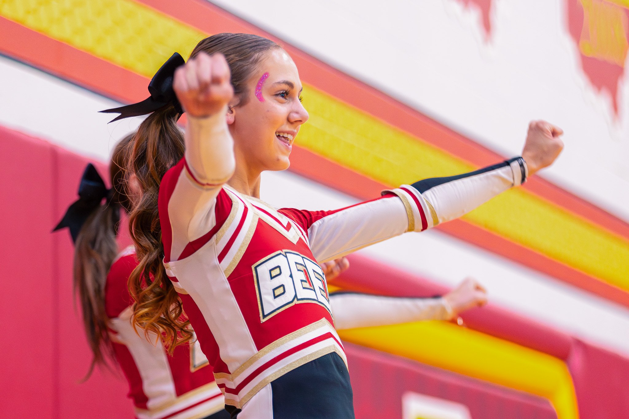 Cheerleader smiling and cheering at a sports event, wearing a red and white uniform with the number 86, and a pink glitter sticker on her cheek.