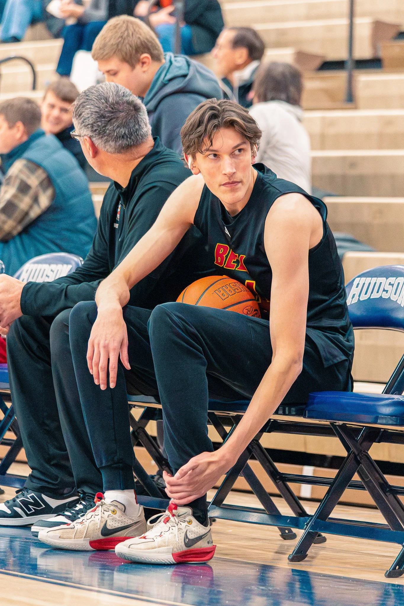 A young male basketball player sitting on a bench holding a basketball, wearing a black sleeveless jersey and athletic shoes, with other players and spectators in the background at an indoor basketball game.