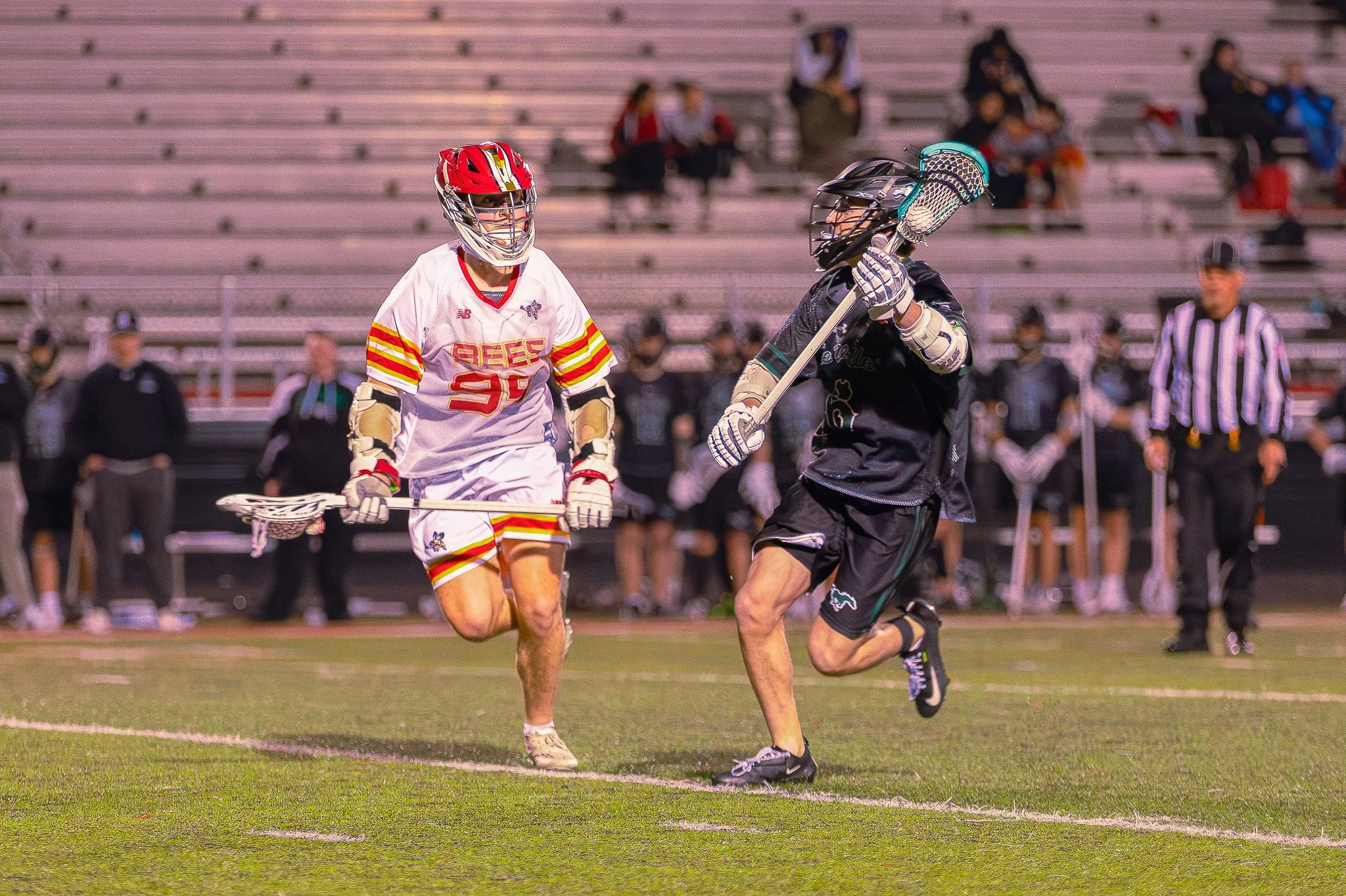 A lacrosse player in a white uniform with red and yellow stripes and a red helmet is running on a field, with another player in black and teal behind him, and spectators in the background.