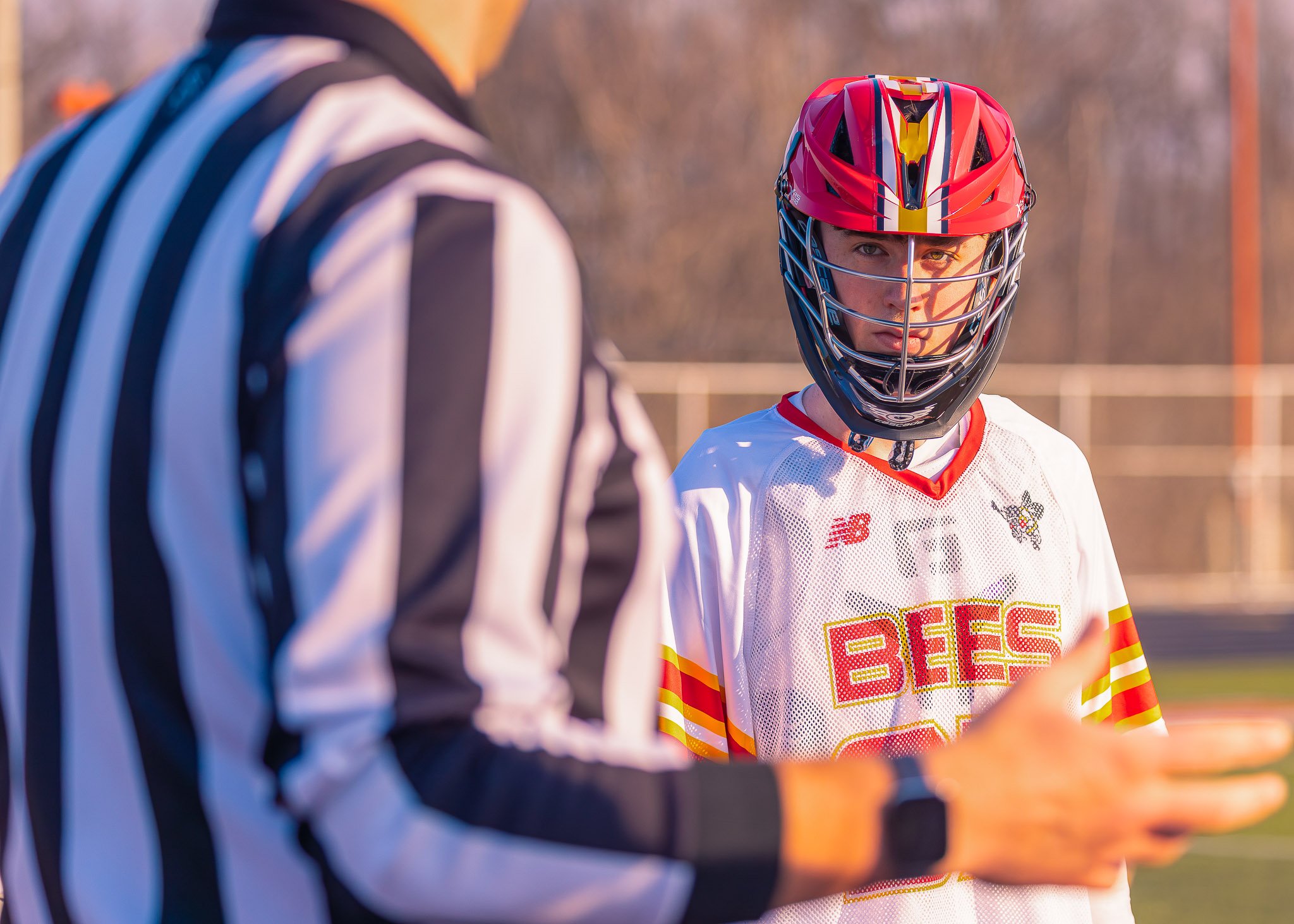 A young male lacrosse player wearing a red helmet with a face cage and a white jersey with red and yellow accents, standing outdoors during late afternoon or early evening, talking to a person in a black and white striped referee uniform.