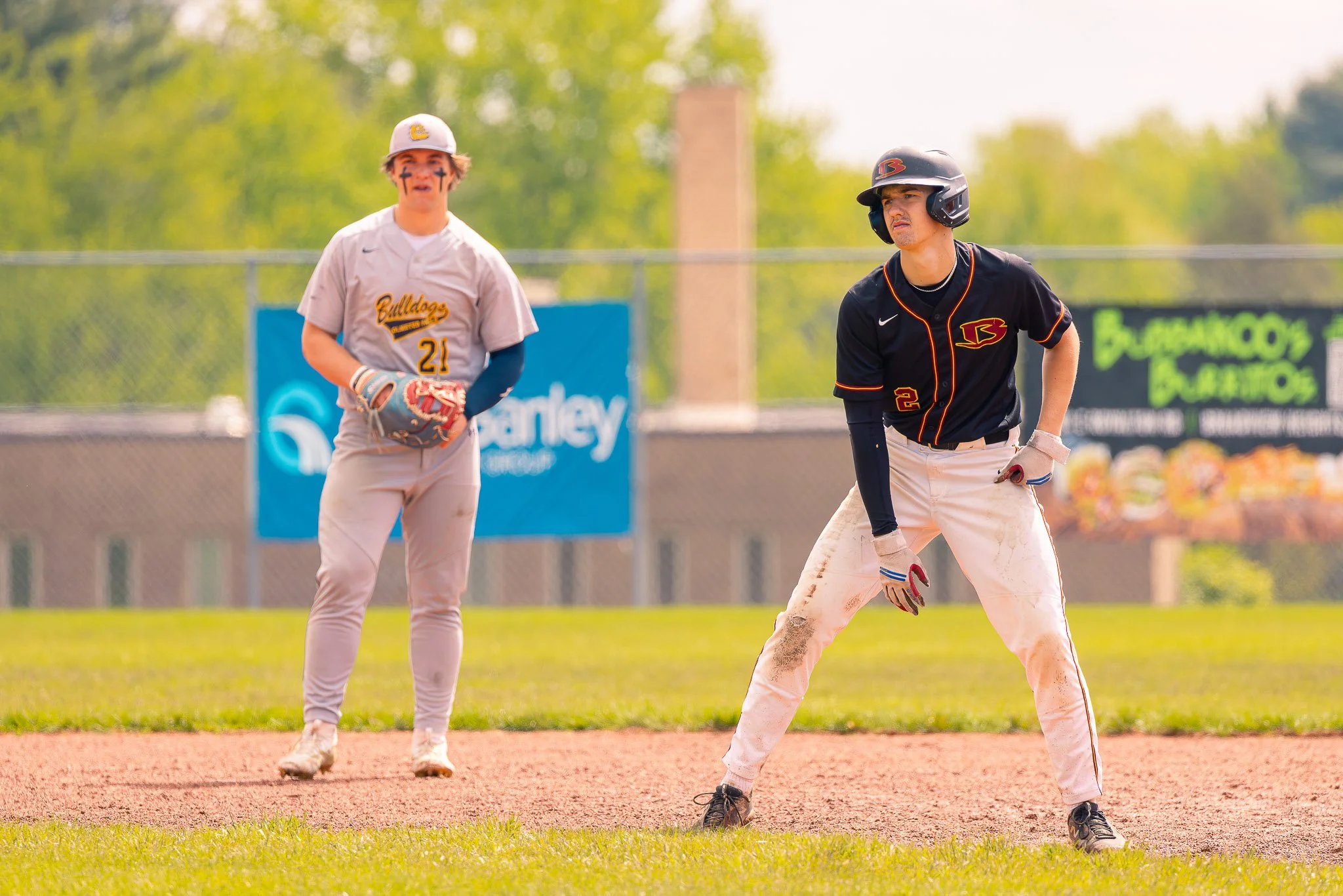 Two baseball players standing on the field with a green background, one in a gray jersey and the other in a black jersey, both wearing helmets and baseball gloves.