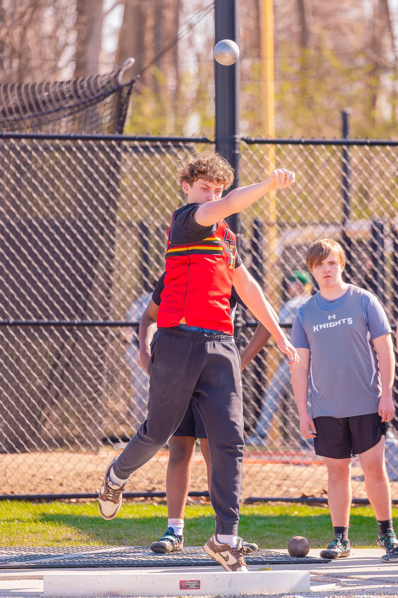 A boy is participating in a shot put event on a field with a fence and trees in the background. He is in the process of releasing the shot put, wearing a red and black sports uniform, and is captured mid-throw.