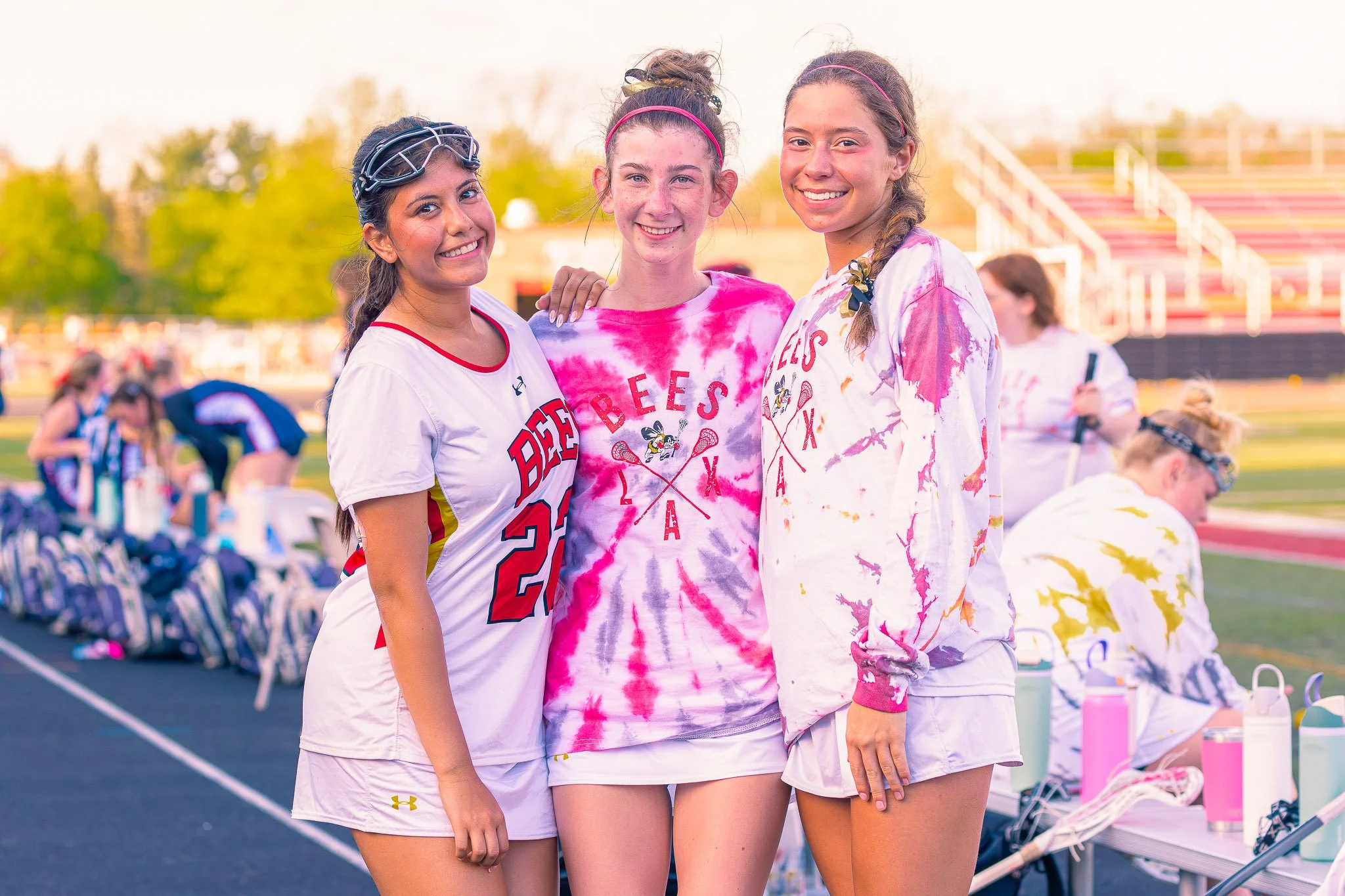 Three young women with athletic build smiling and standing close together outdoors on a sports field, wearing sportswear and tie-dye shirts, with other people and bleachers in the background.