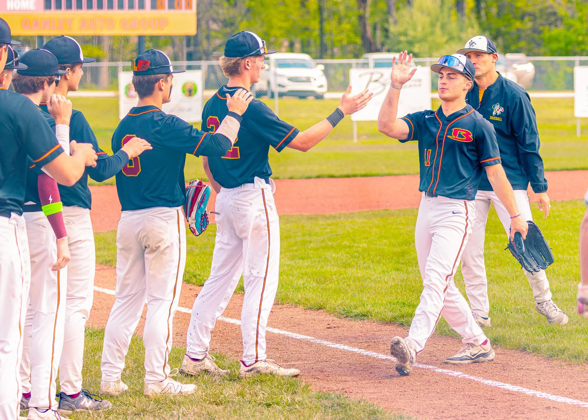 A group of young baseball players on a field, engaging in a team high-five before a game. They are wearing navy blue jerseys with orange and yellow accents, white pants, and caps. Some are wearing sunglasses or wristbands.