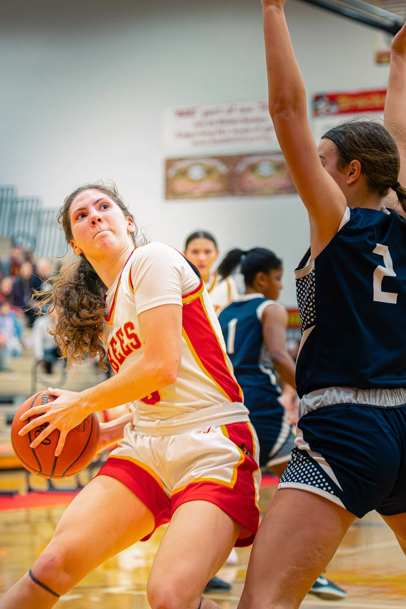 Two female basketball players playing in a gymnasium. One wearing a white and red uniform holding a basketball, and the other wearing a dark blue uniform defending. Spectators and a scoreboard are visible in the background.