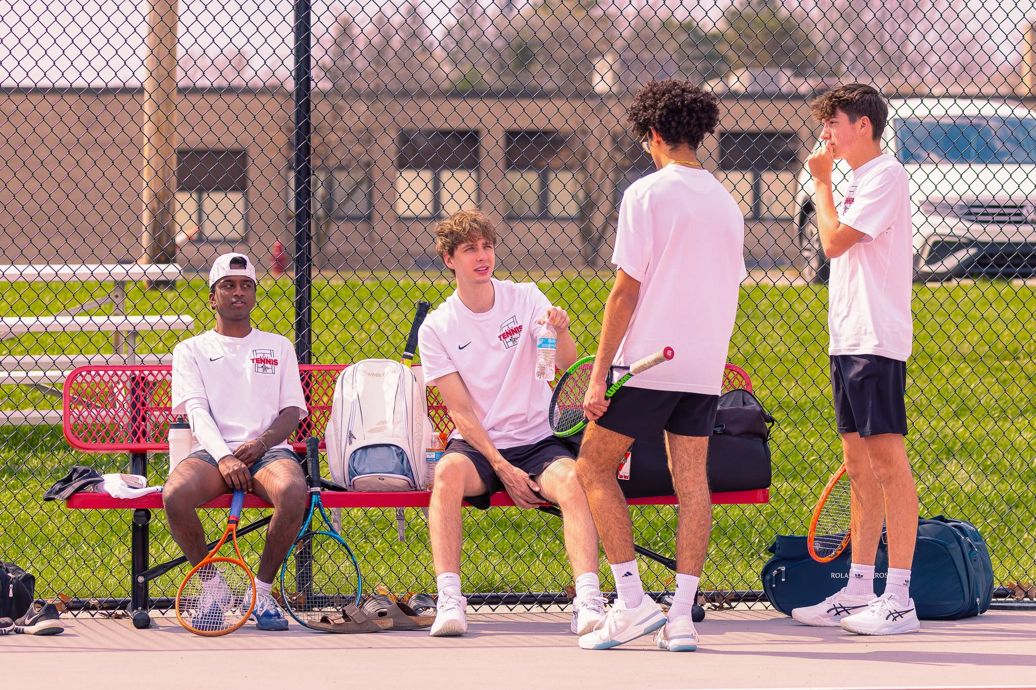 Four young male tennis players on a tennis court, sitting and standing near a chain-link fence, with tennis racquets, bags, and water bottles nearby, preparing for a match or practice.