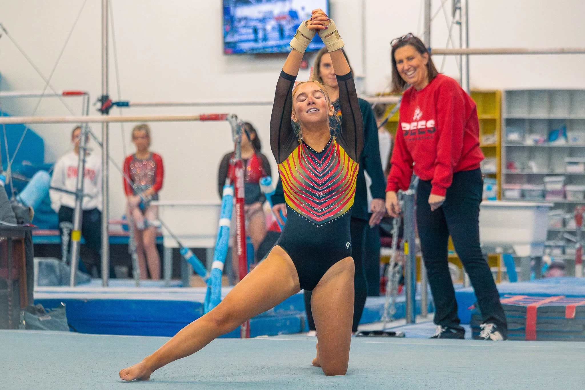 A female gymnast in a colorful leotard is performing a split on the gym floor, with her eyes closed and hands clasped above her head. Several women, some wearing red team sweatshirts, are watching and smiling in the background at a gymnastics trainin