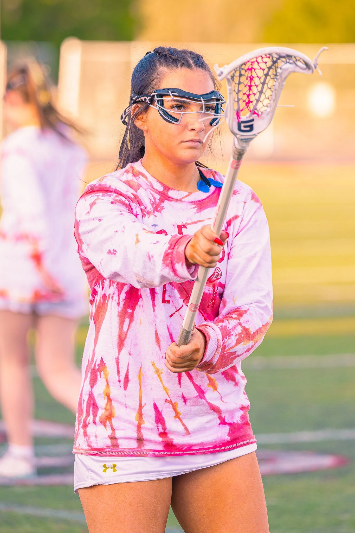 A young female lacrosse player in a pink and white tie-dye long sleeve shirt and white shorts, holding a lacrosse stick, on a field during daytime. She is wearing protective goggles.