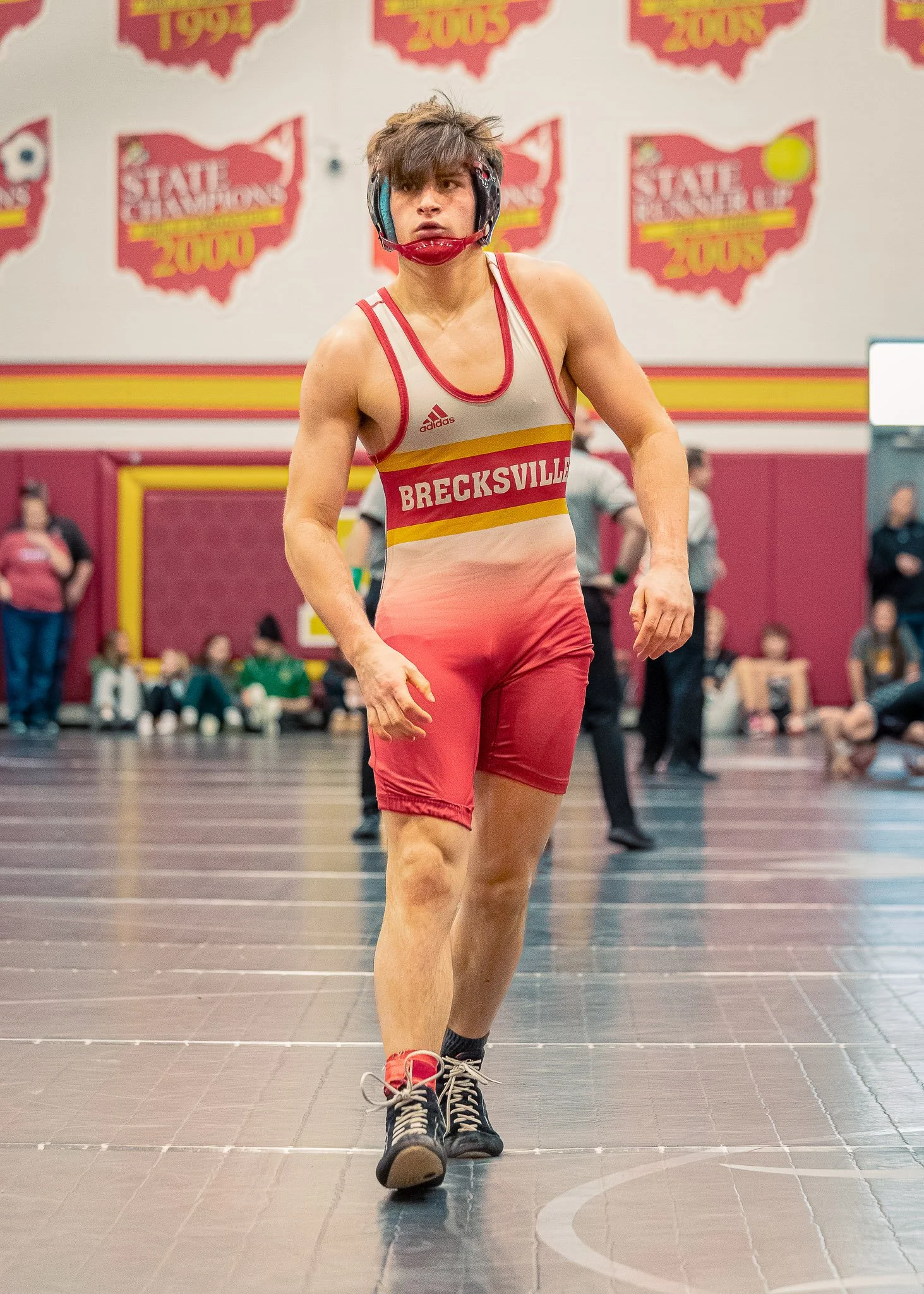 Young male wrestler wearing a red and beige singlet with 'BRECKSVILLE' printed on it, black and red wrestling shoes, and a helmet, walking on a wrestling mat in a gym with banners on the wall.