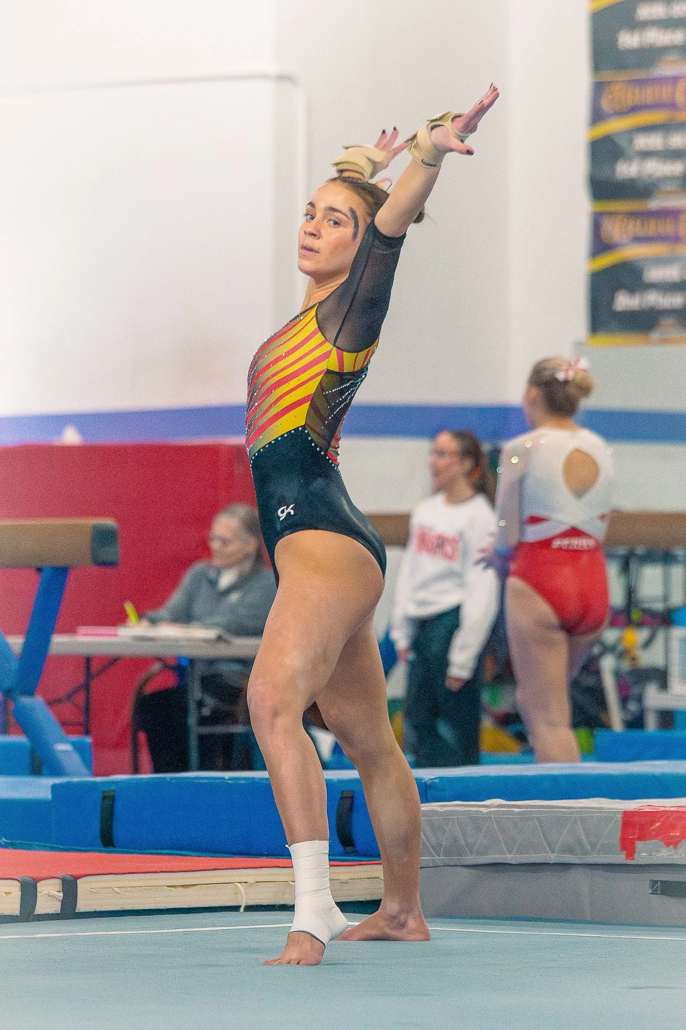 A female gymnast standing, wearing a colorful leotard with black mesh sleeves and white socks, with her arms raised above her head. Other gymnasts and officials can be seen in the background during a gymnastics competition.