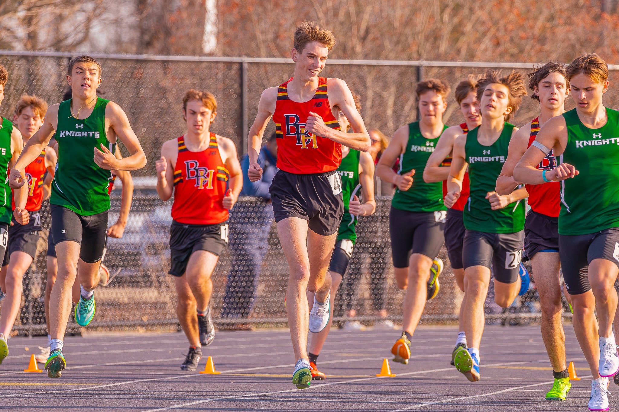 Group of teenagers running in a cross-country race on an outdoor track, some wearing green and others in red and yellow uniforms.