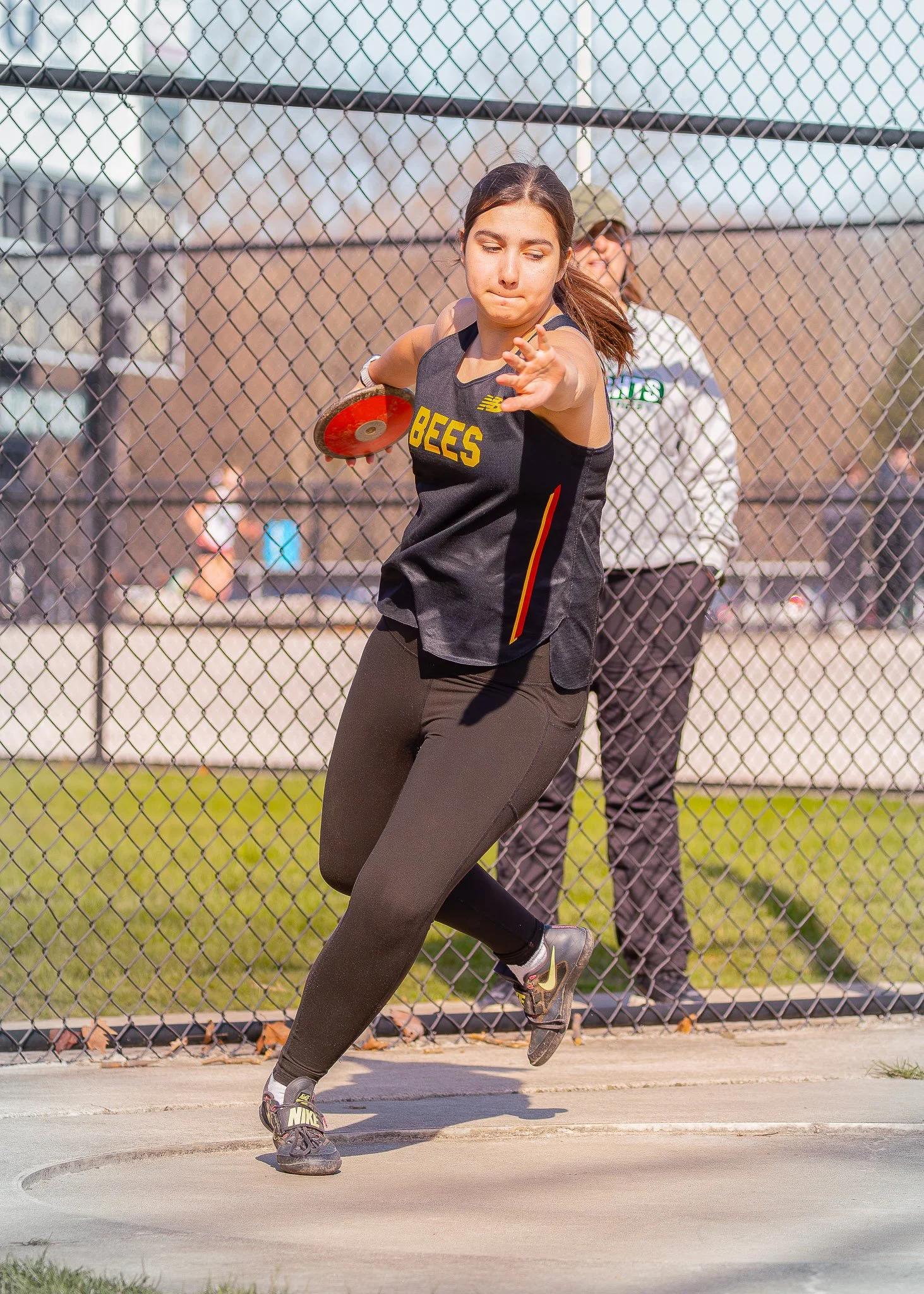 A female athlete in a black Bees sports jersey and black athletic pants is throwing a discus on a track and field area, with a chain-link fence behind her and a person in a white shirt and glasses in the background.