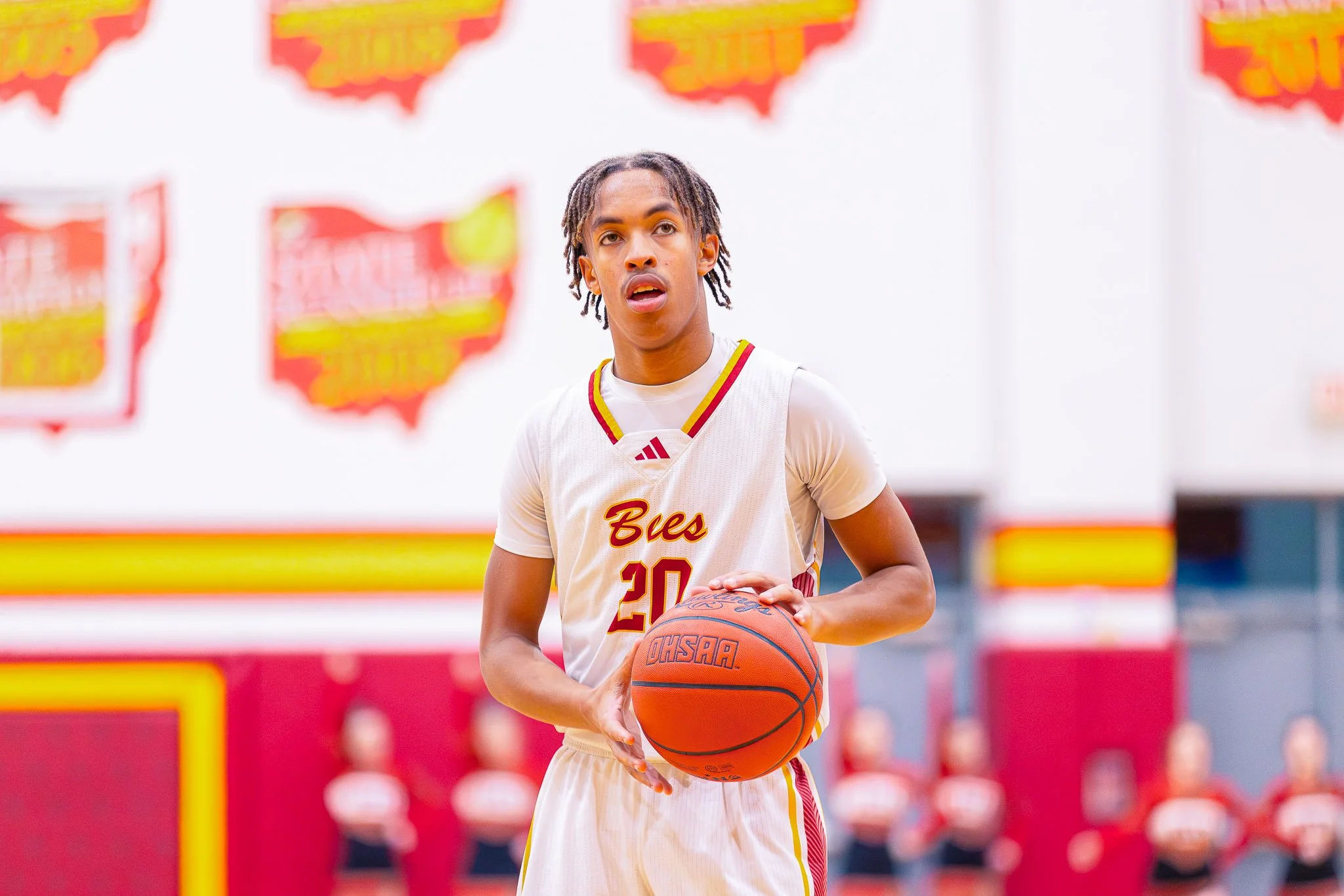 A young male basketball player wearing a white jersey with red and yellow accents, holding a basketball and looking towards the side, on a basketball court with branded backboards and red and yellow wall padding in the background.
