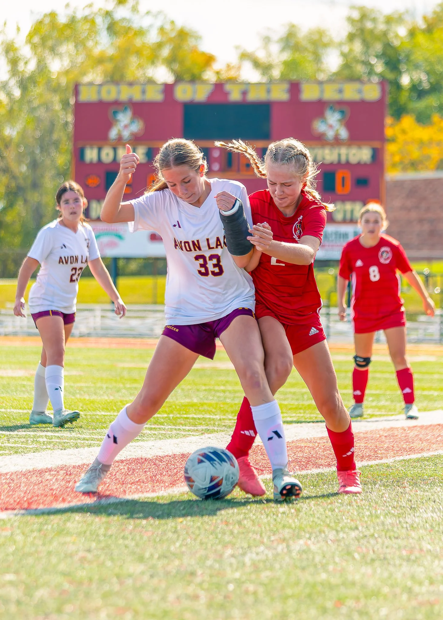 Two young female soccer players are competing for the ball on the field, with other players and a scoreboard in the background.