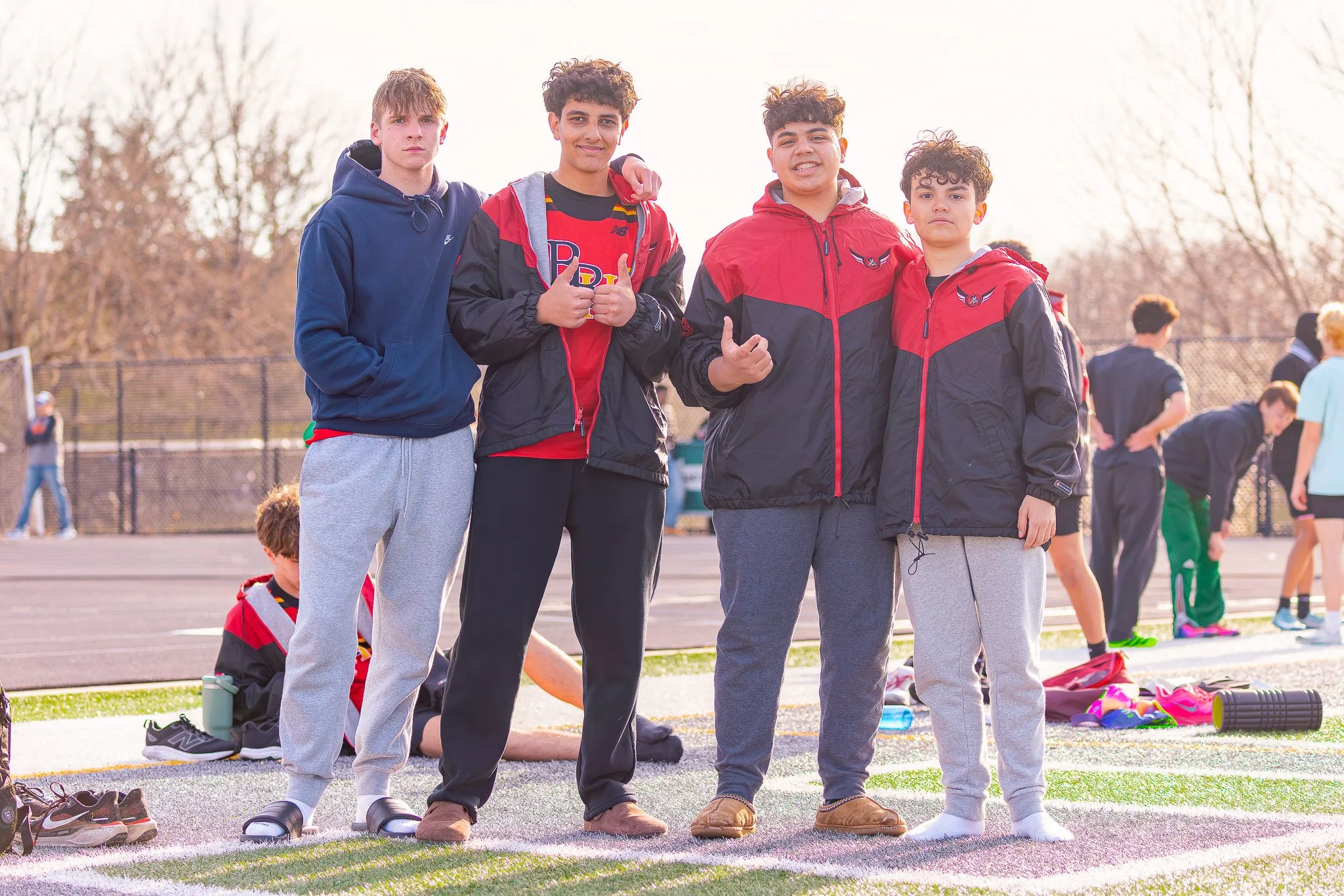 Four boys standing together on an outdoor sports field, with other children in the background, during daytime.