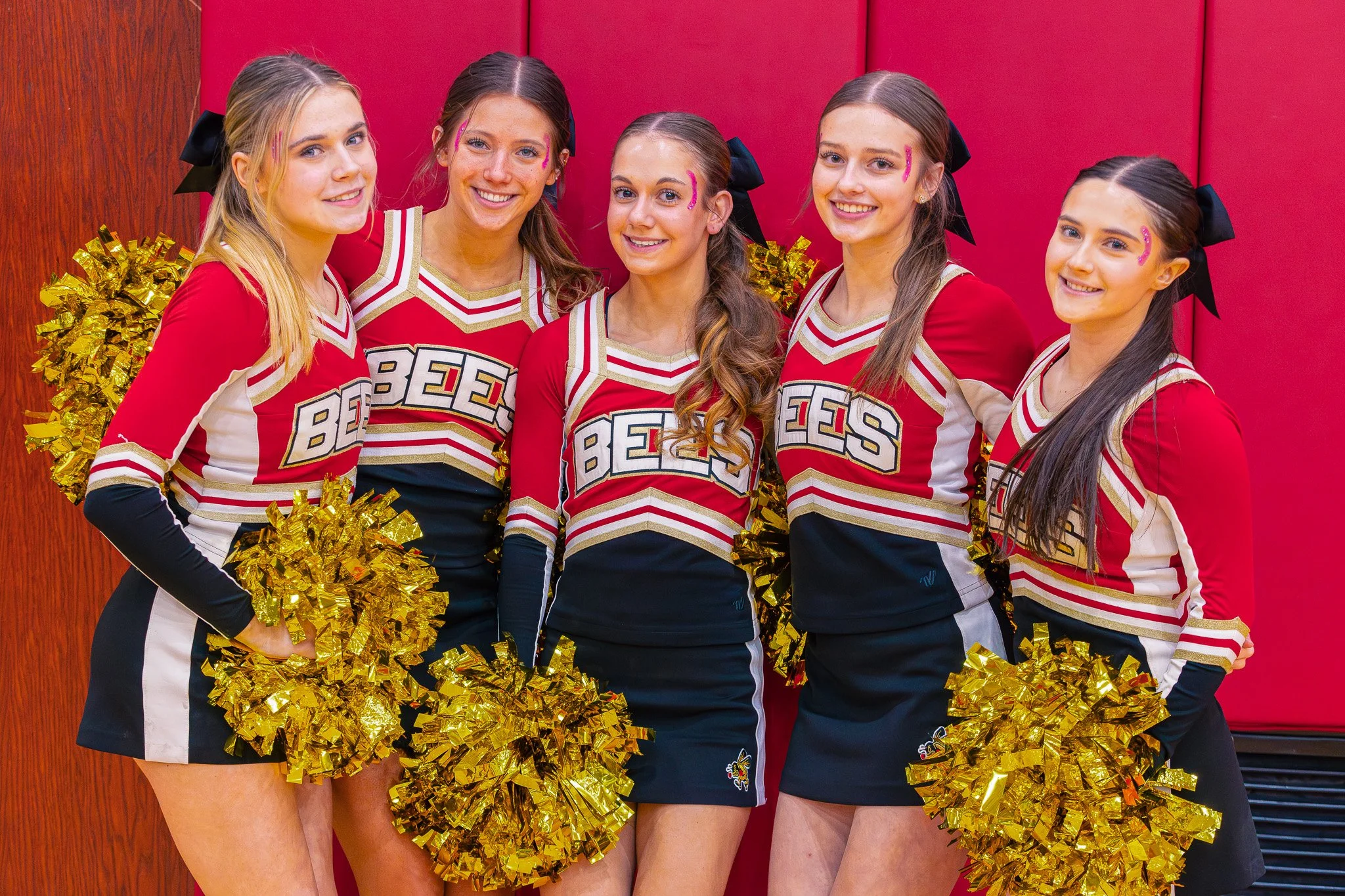Group of five cheerleaders in red and white uniforms holding gold pom-poms, standing in front of a red padded wall.