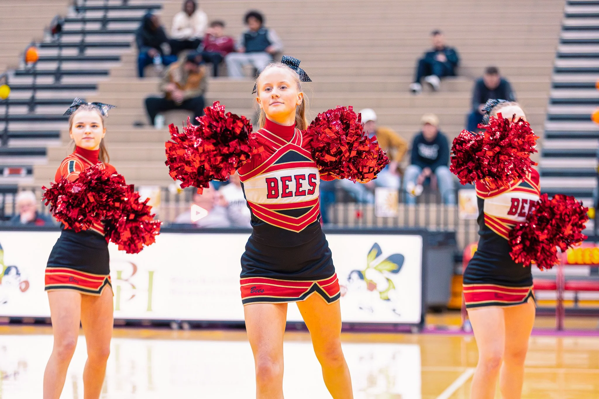 Young female cheerleader in a red, black, and gold uniform holding red pom-poms in a gymnasium during a cheerleading routine, with other cheerleaders in similar uniforms, and spectators sitting on bleachers in the background.