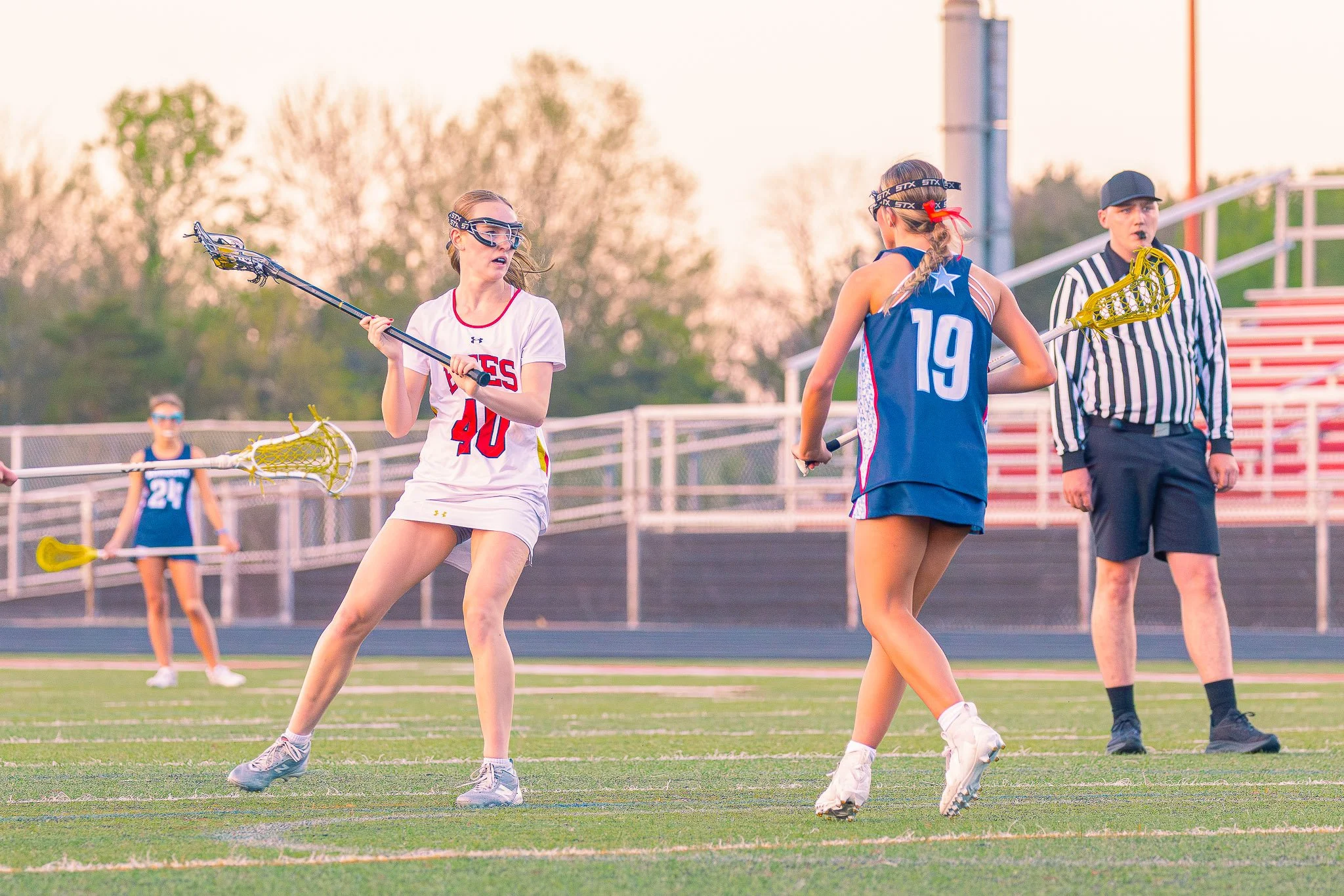 Girls playing lacrosse on a field during sunset, with a referee nearby.