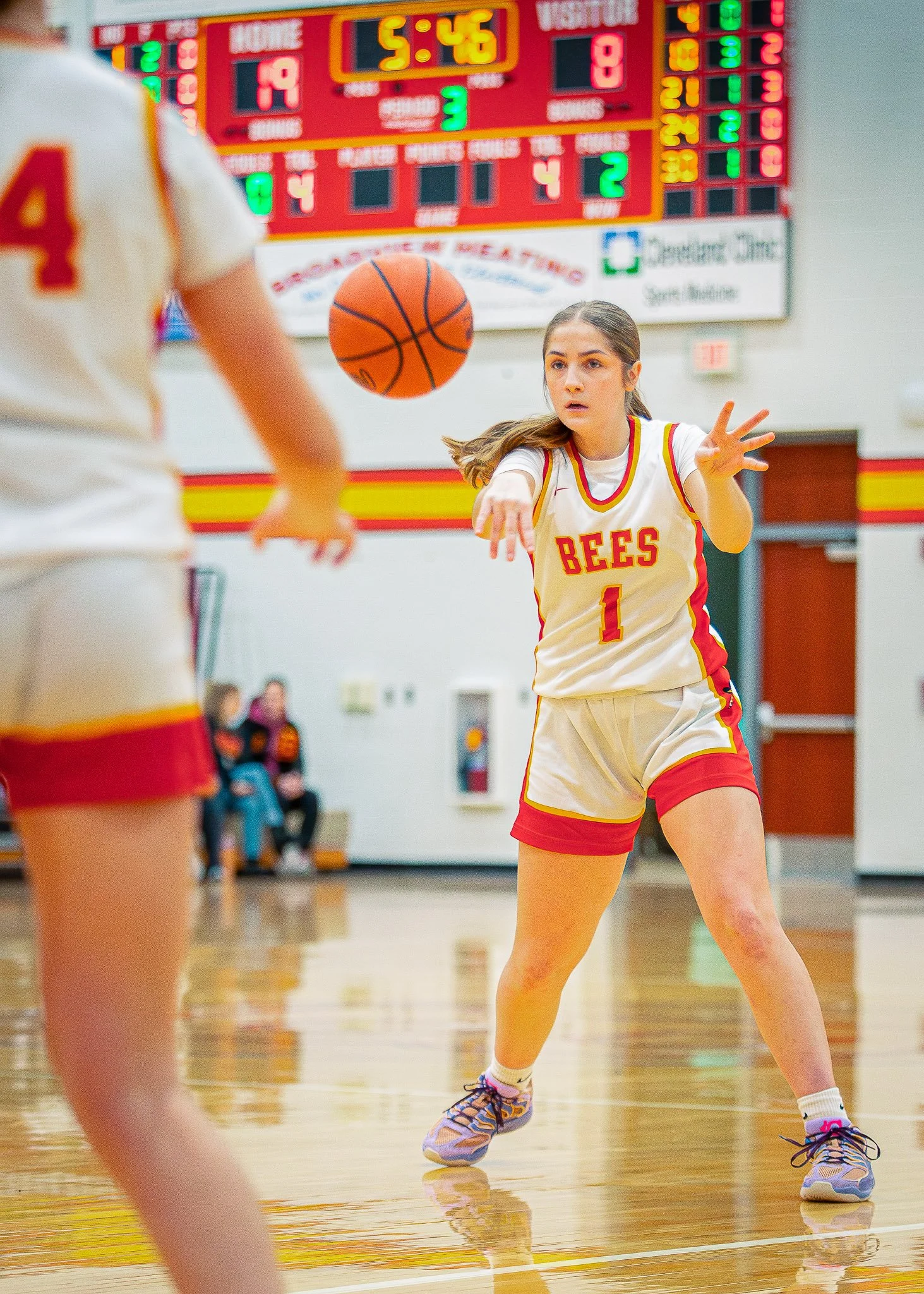 A girl wearing a yellow and red basketball uniform with the number 1 on it, playing basketball on an indoor court, with a scoreboard and spectators in the background.