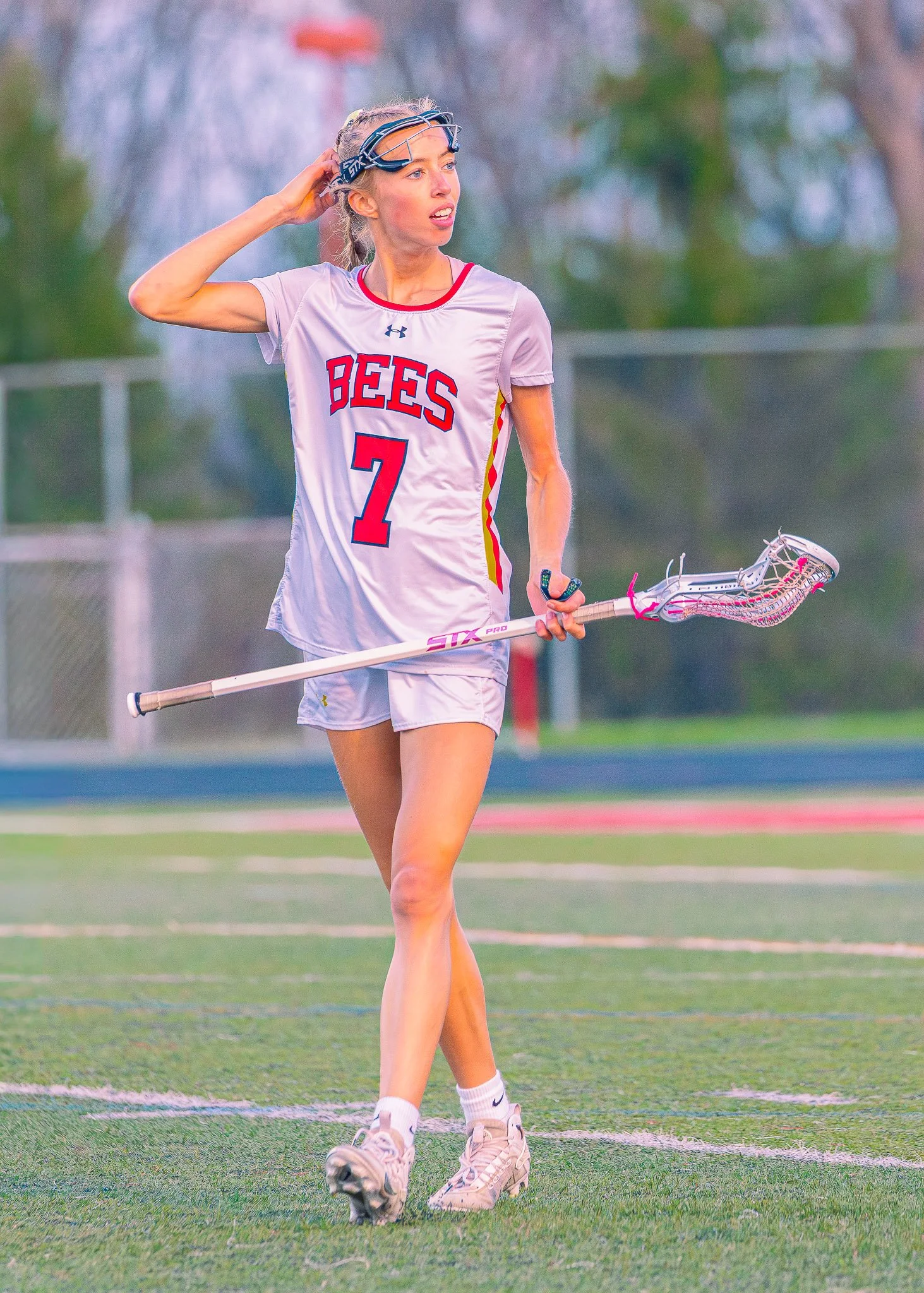 A female lacrosse player on a field, wearing a white jersey with 'BEES' written on it and the number 7, holding a lacrosse stick, with a protective headgear pushed up on her forehead, and an expression of surprise or concern on her face.