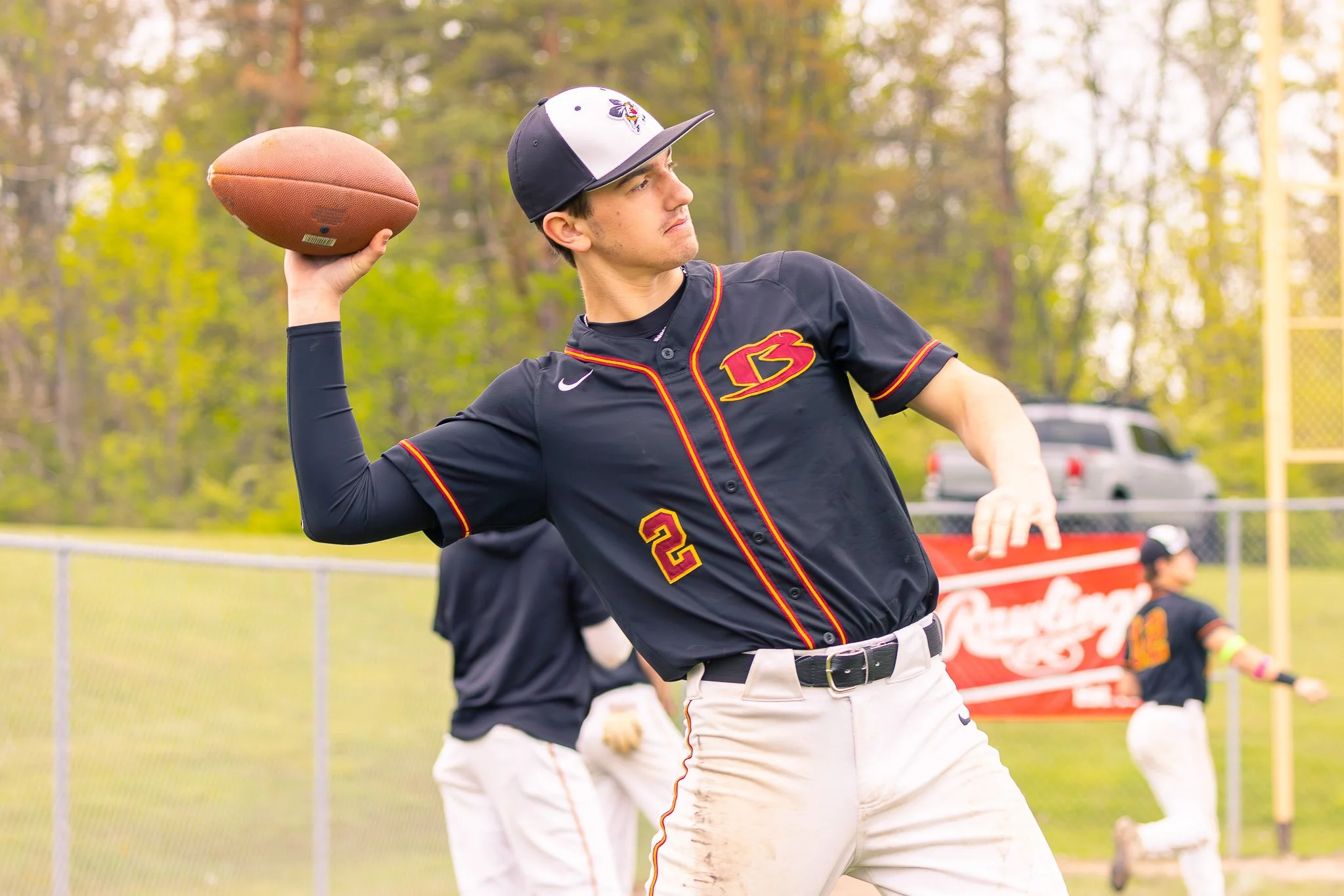 Young man wearing a black and yellow baseball uniform with the number 2, holding a football in his right hand, preparing to throw, on a baseball field with trees in the background