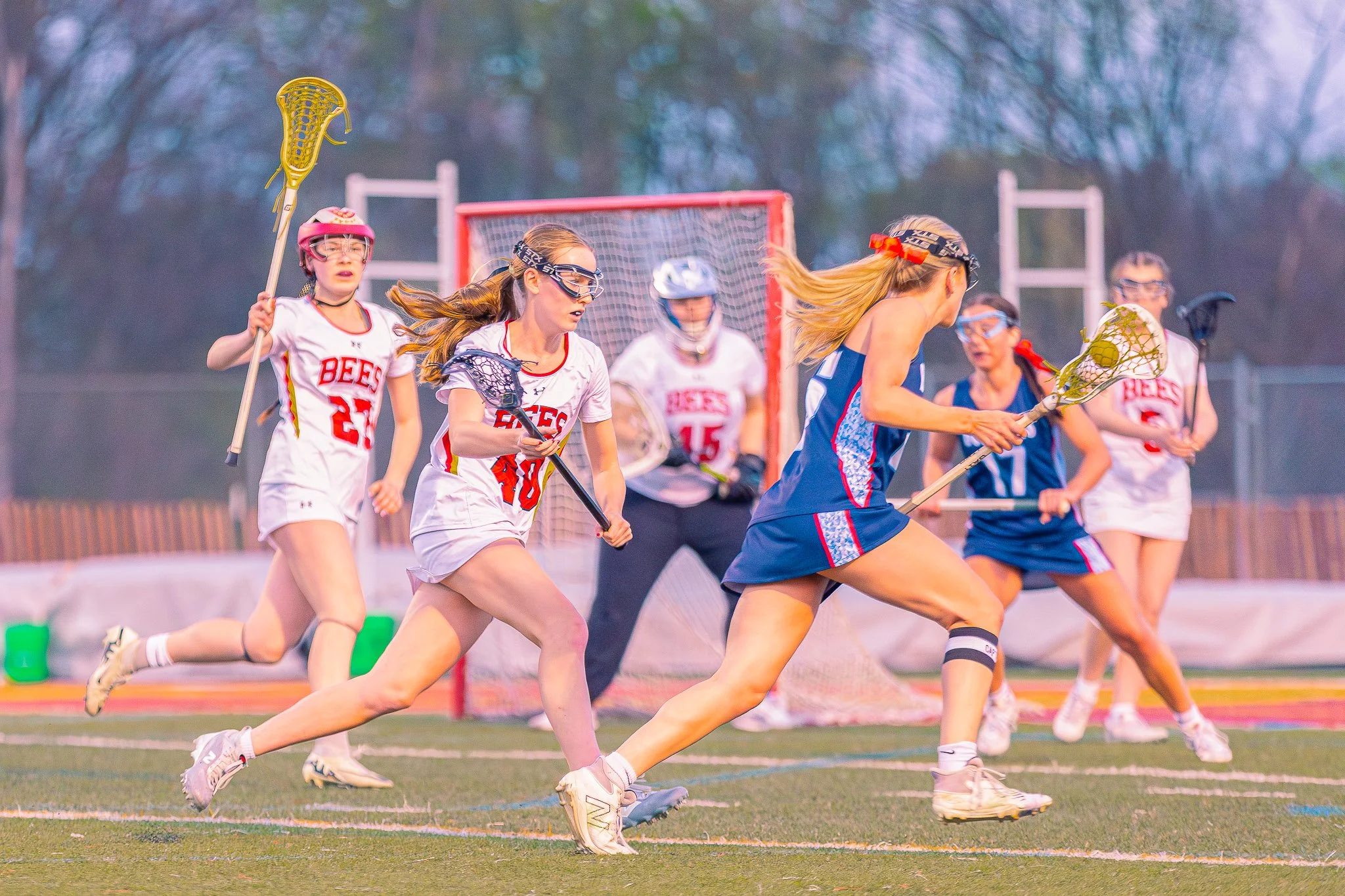 Girls playing lacrosse on a field during a game, with a goalpost and trees in the background.