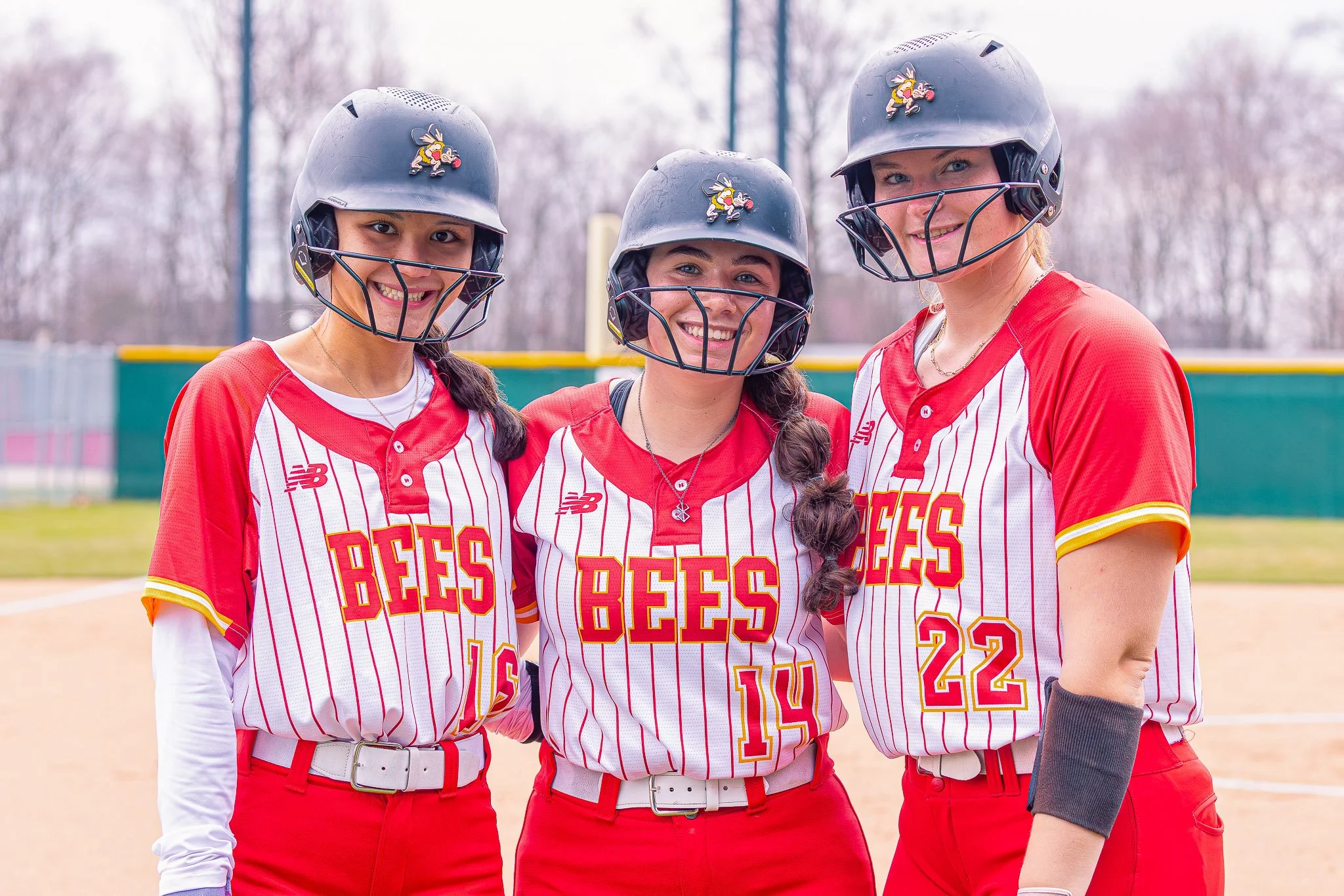Three female softball players standing together on a field, wearing gray helmets with a bee mascot, red and white striped uniforms with the text "BEES," and red pants, smiling at the camera.