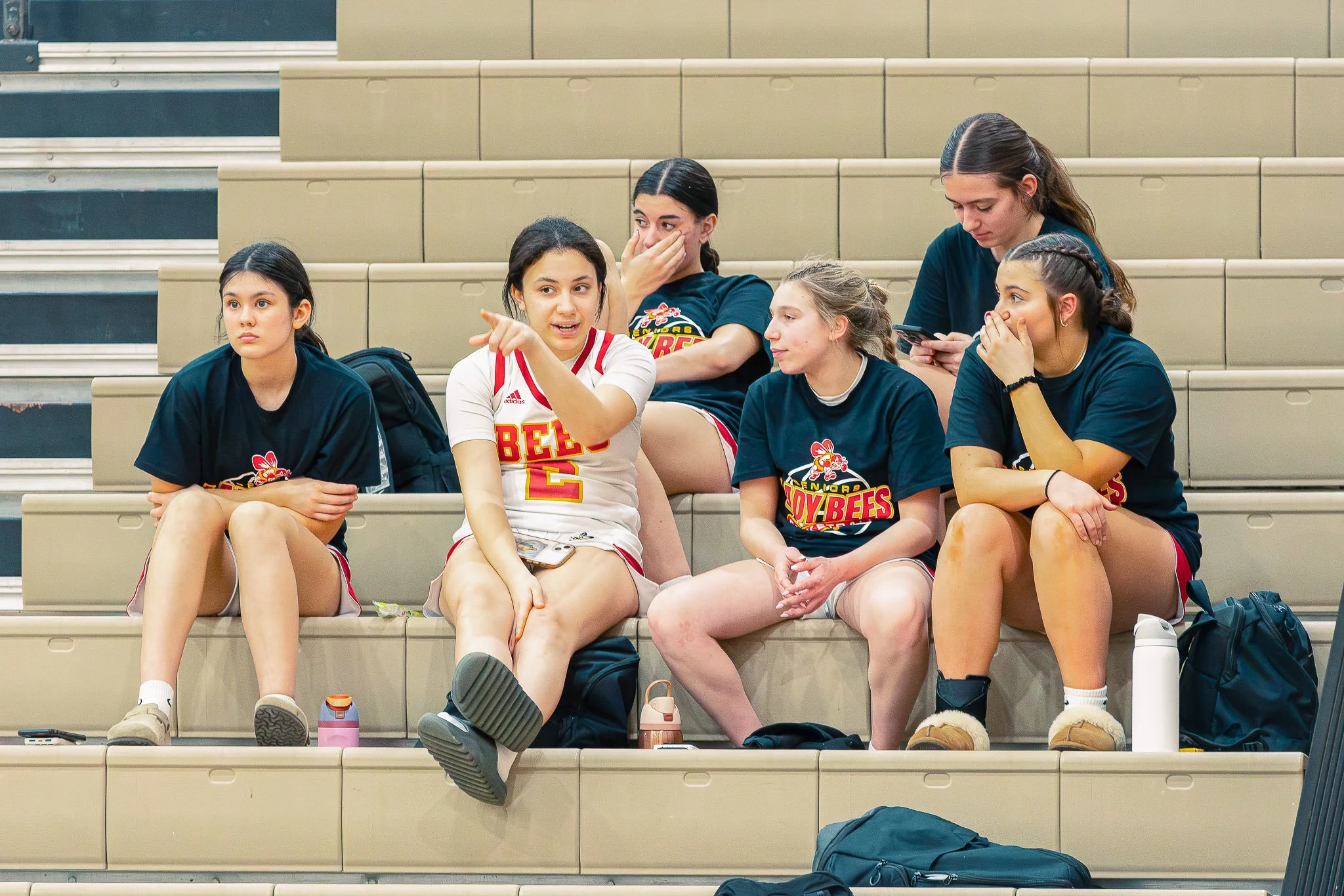 Five young women sitting on bleachers in a gymnasium, wearing basketball uniforms, some with sports drinks and backpacks around them, engaged in conversation or using their phones.