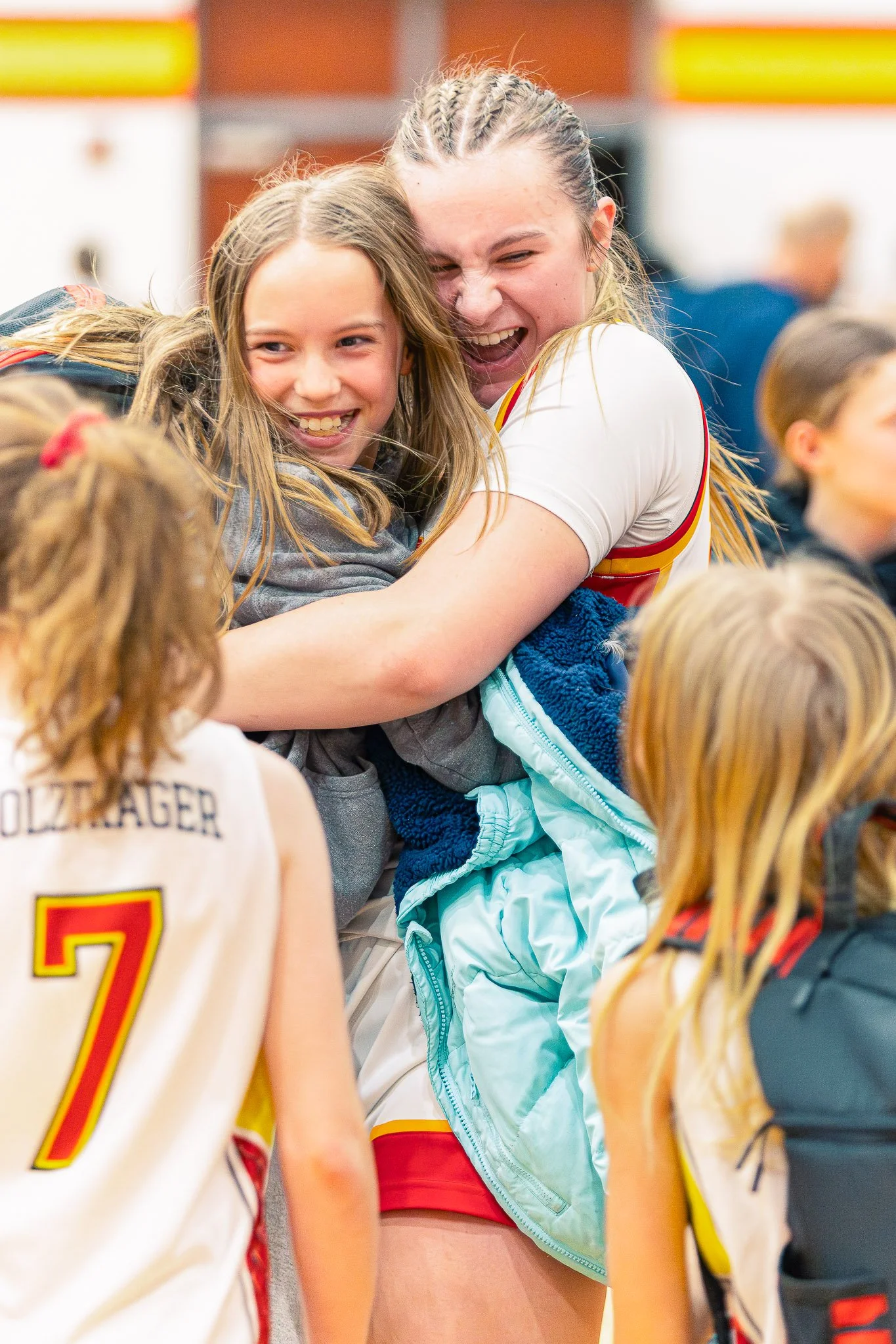 Women hugging and smiling at a sports event with children around them, wearing basketball uniforms.