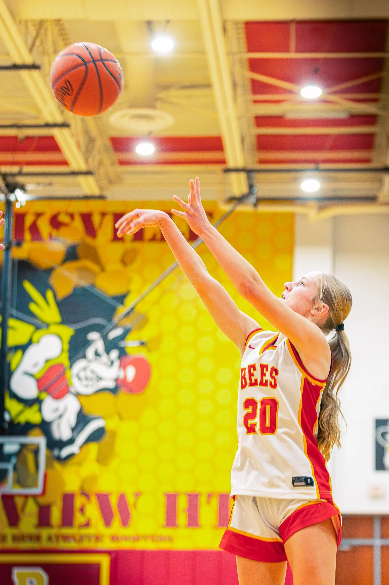 A young female basketball player in a white and red uniform with the number 20 is playing basketball indoors, reaching to shoot or catch the ball.
