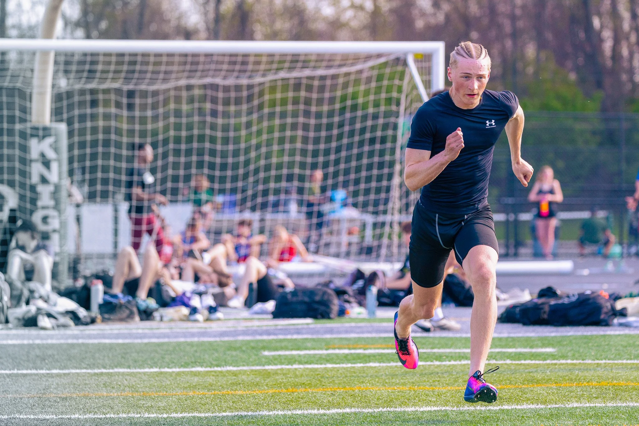 A male athlete running on a track during an outdoor sports event, with spectators sitting on benches behind a goalpost and trees in the background.