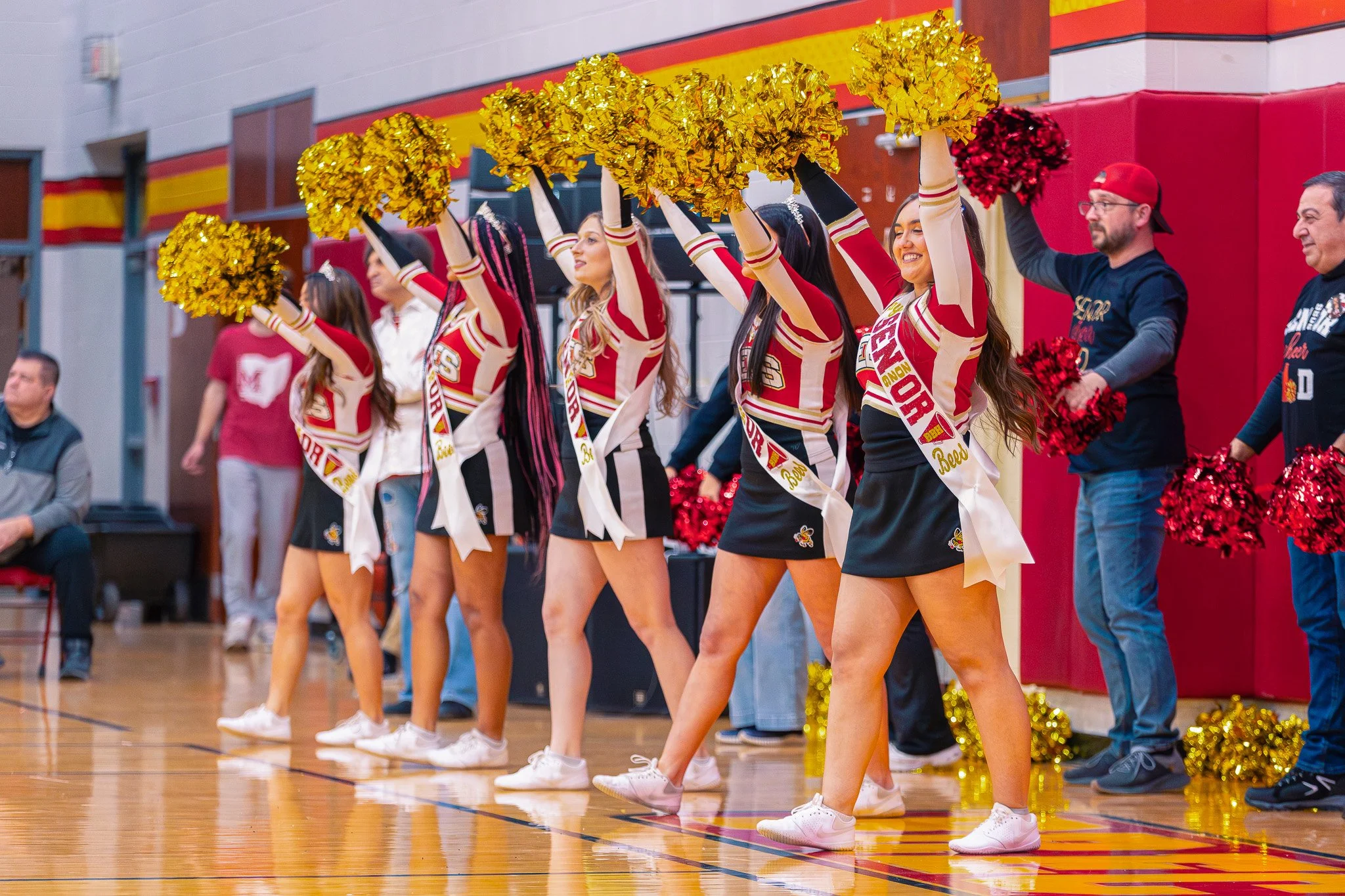 Cheerleaders performing a routine on the gymnasium floor during a sports event, wearing red and black uniforms and holding gold pom-poms.