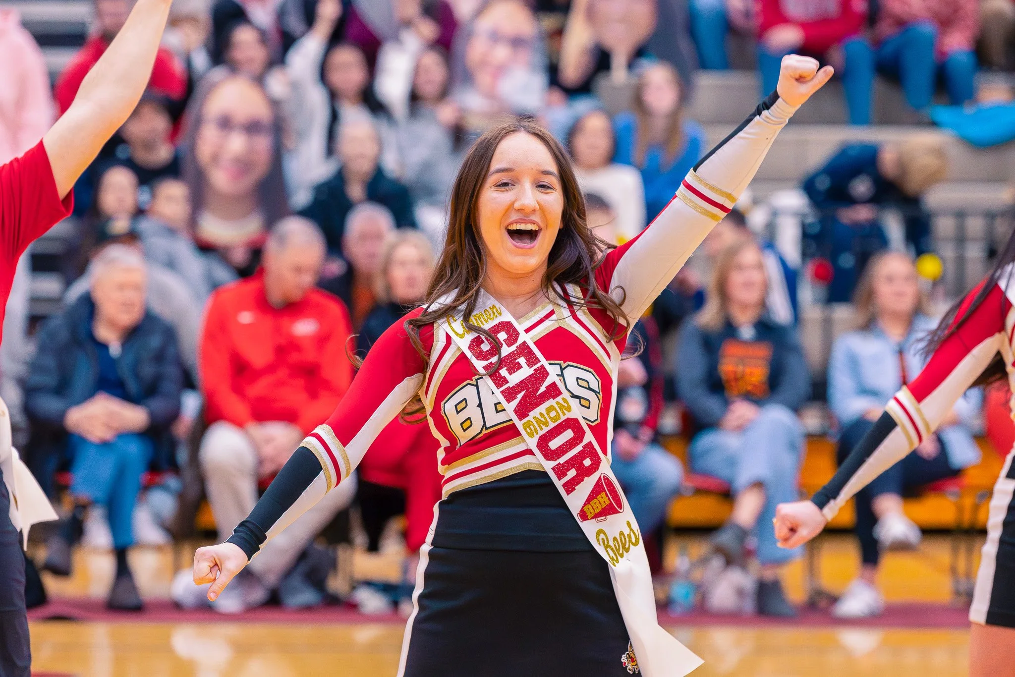 Smiling cheerleader in a red, white, and black cheerleading uniform raising one arm in a cheer at a sports event with a crowd in the background.