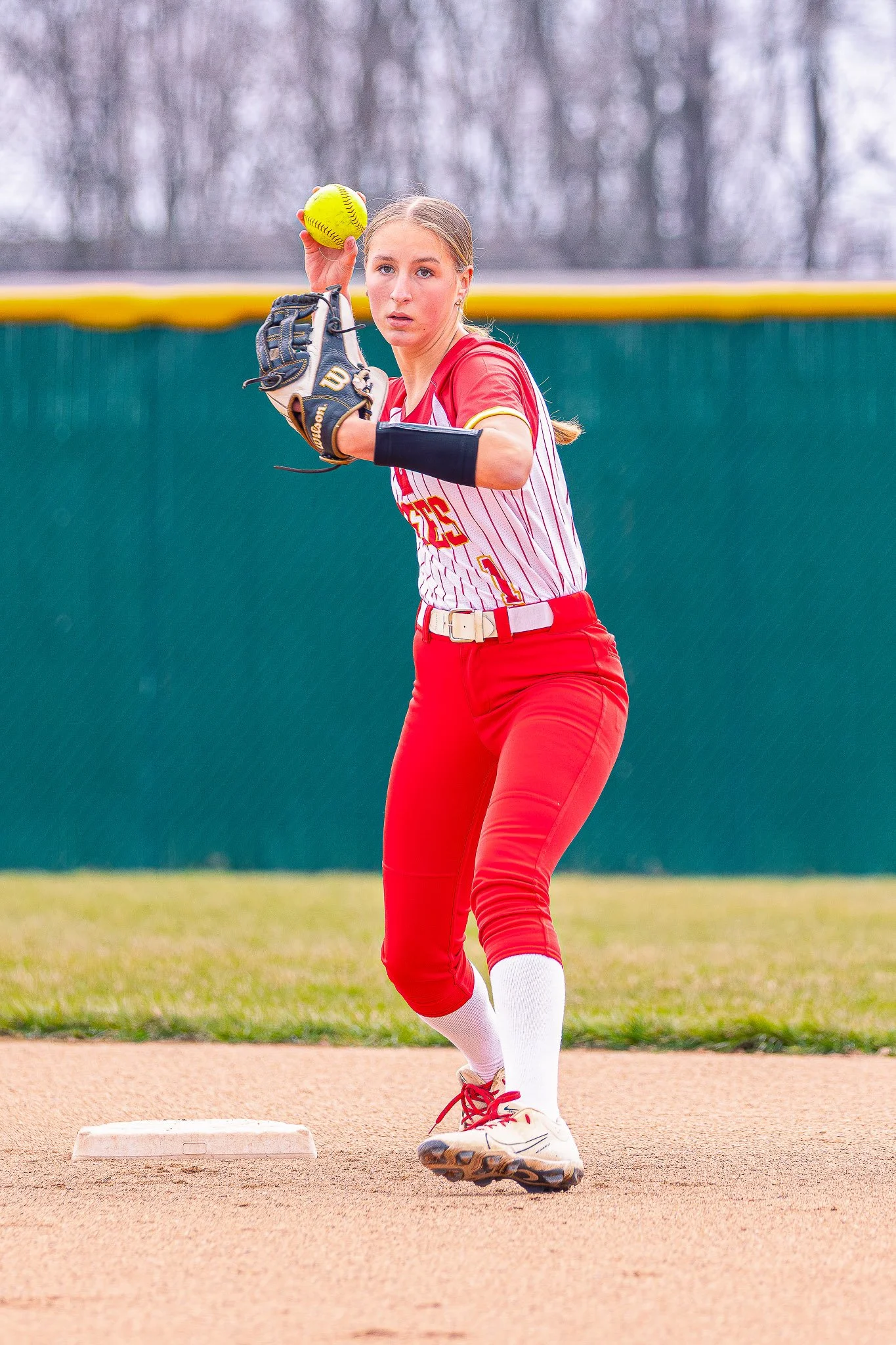 A female softball player on the field, holding a yellow softball above her head, ready to pitch, wearing a red and white uniform, and standing near the base on a dirt field with green outfield in the background.