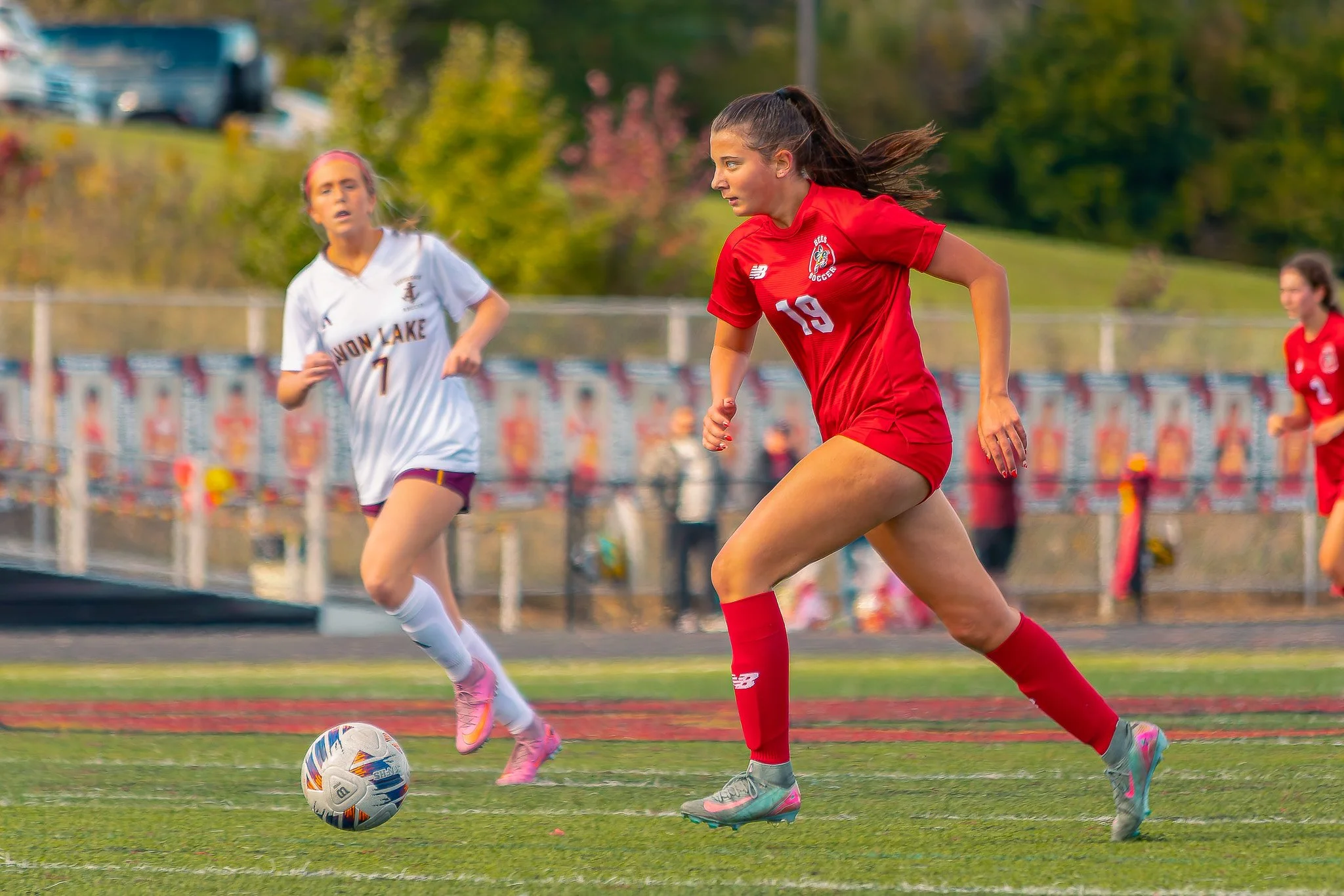Two female soccer players, one in a red uniform and the other in a white uniform, compete for the ball on a soccer field during a game, with trees and cars in the background.
