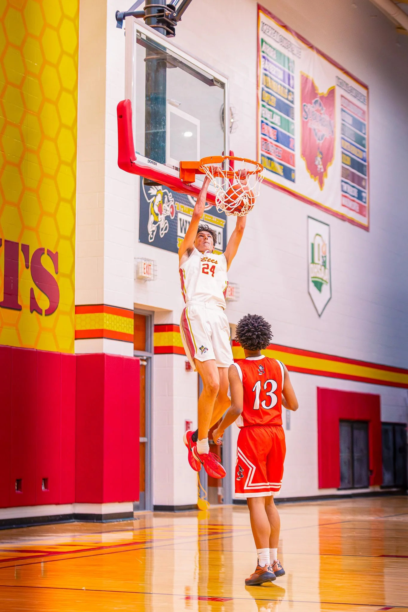A basketball player wearing a white jersey with the number 24 is attempting a dunk at the basketball hoop while another player wearing a red jersey with the number 13 is watching from below in a gymnasium.