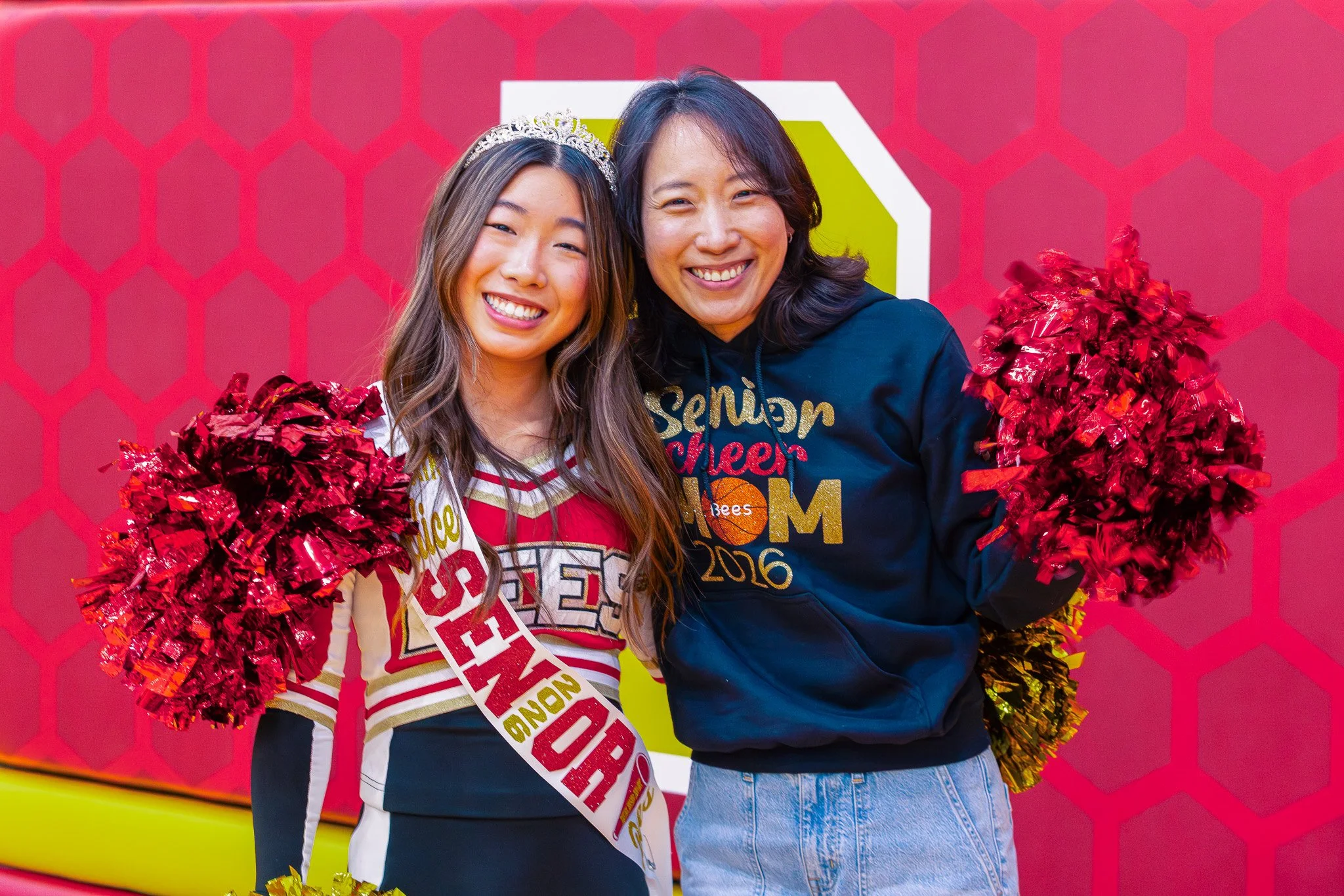 Two women smiling, holding red and gold pom-poms, one wearing a tiara and a sash that says 'Senior', the other wearing a navy hoodie with gold text, standing in front of a pink background with a honeycomb pattern.