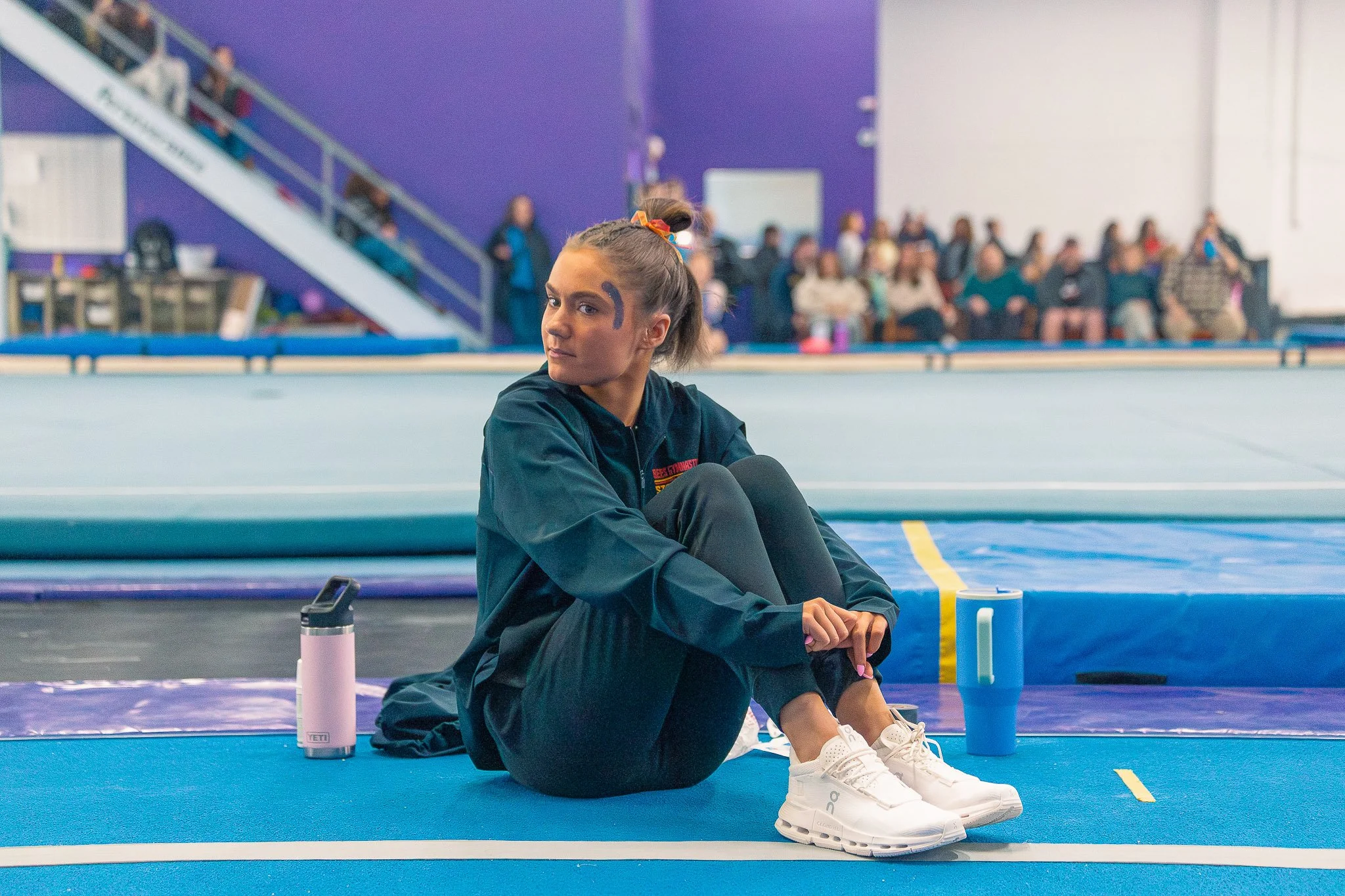 A young female gymnast sitting on a blue exercise mat in a gymnasium, wearing a black jacket and white sneakers, with water bottles nearby. In the background, there are spectators sitting on benches and watching her.