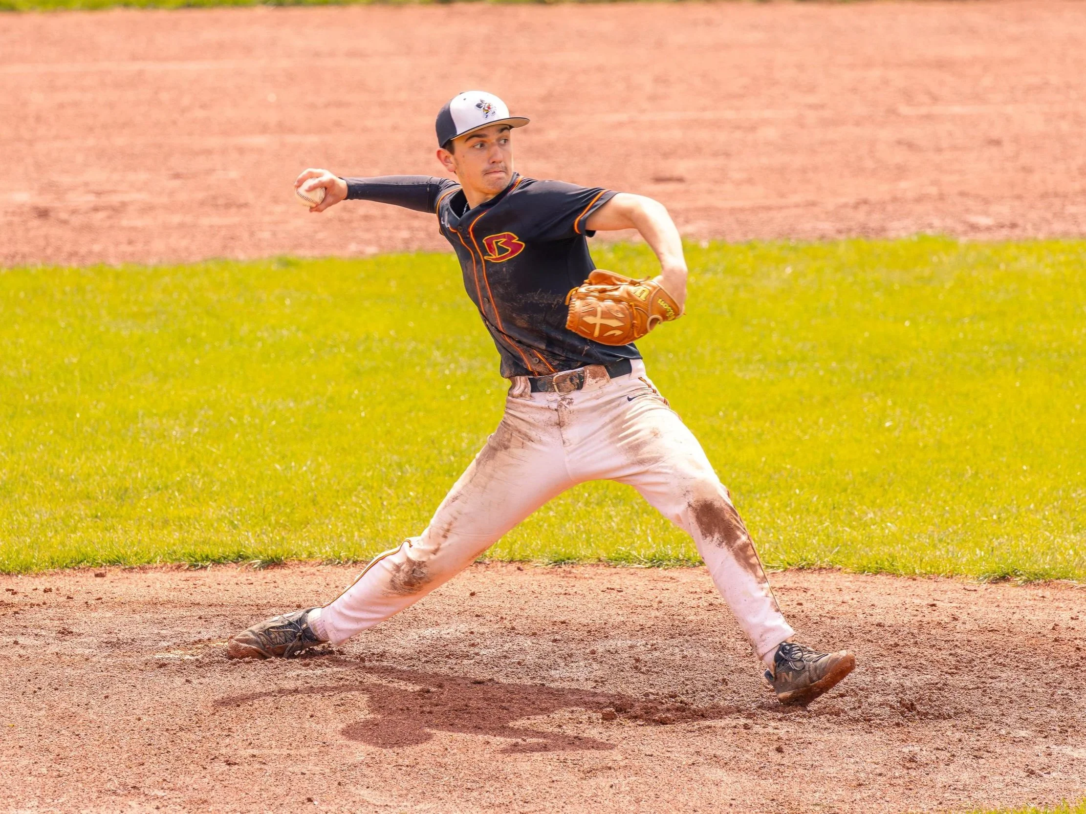 A baseball player in a black jersey and white pants is throwing a baseball on a dirt field with grass and a dirt wall in the background.