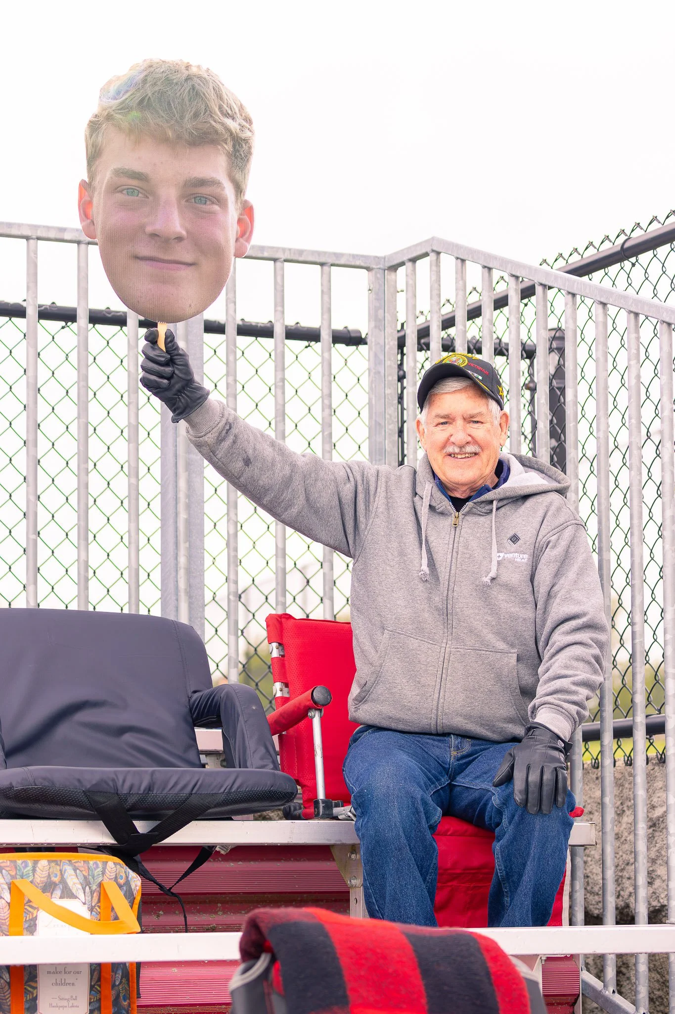 A man with gray hair and a gray hoodie sitting on a red bench, holding a large cutout of a young man's face in his right hand, smiling. There are chairs, bags, and a fence in the background.
