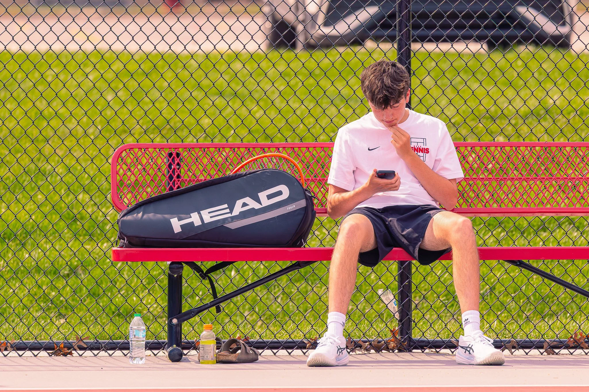 Teenage boy sitting on a pink bench behind a chain-link fence, looking down at his phone, with tennis equipment and water bottles around him on a tennis court.