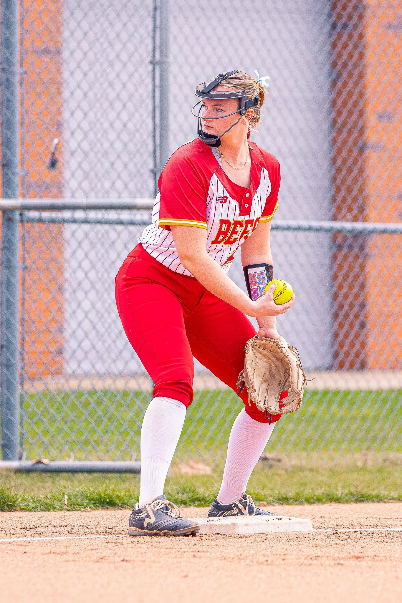 A female softball player in a red and white uniform is preparing to catch a yellow softball with a glove on her left hand at a softball field.