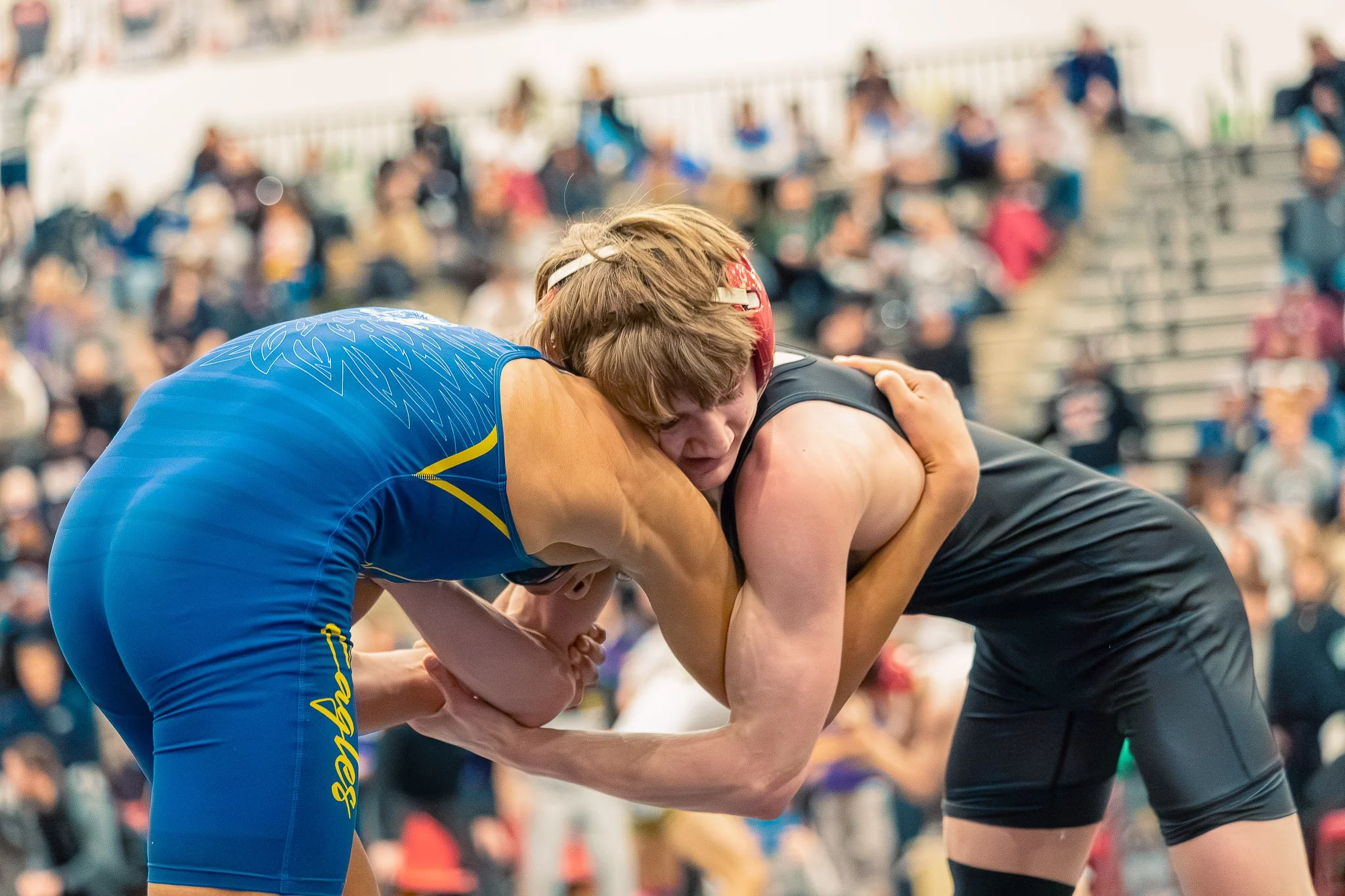 Two female wrestlers in a match, grappling on the mat, with a crowd of spectators watching in the background.