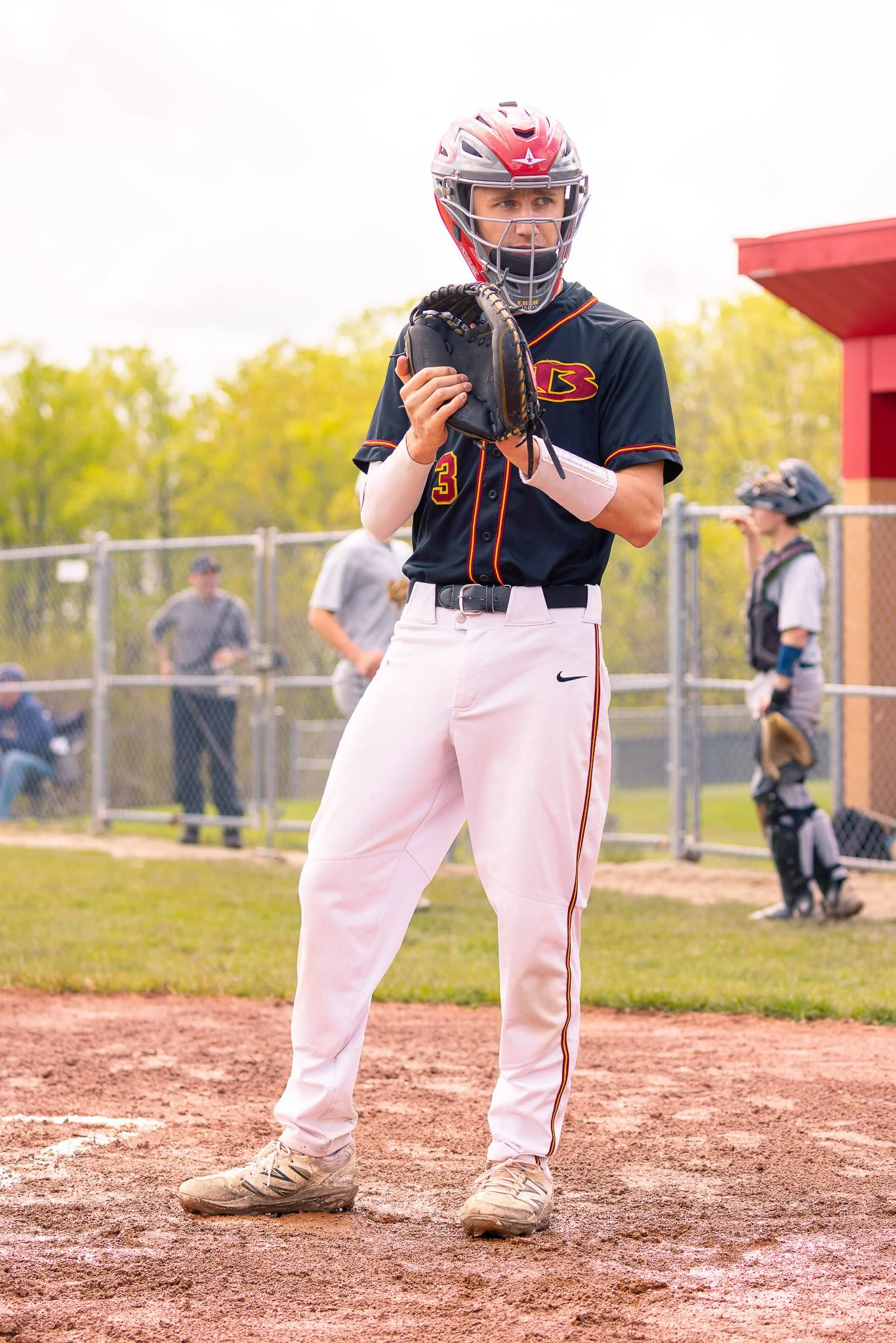 A baseball player in a black jersey with the number 3, standing at home plate on a baseball field. The player is holding a baseball glove and wearing a helmet with a face guard. There are other players and people in the background.