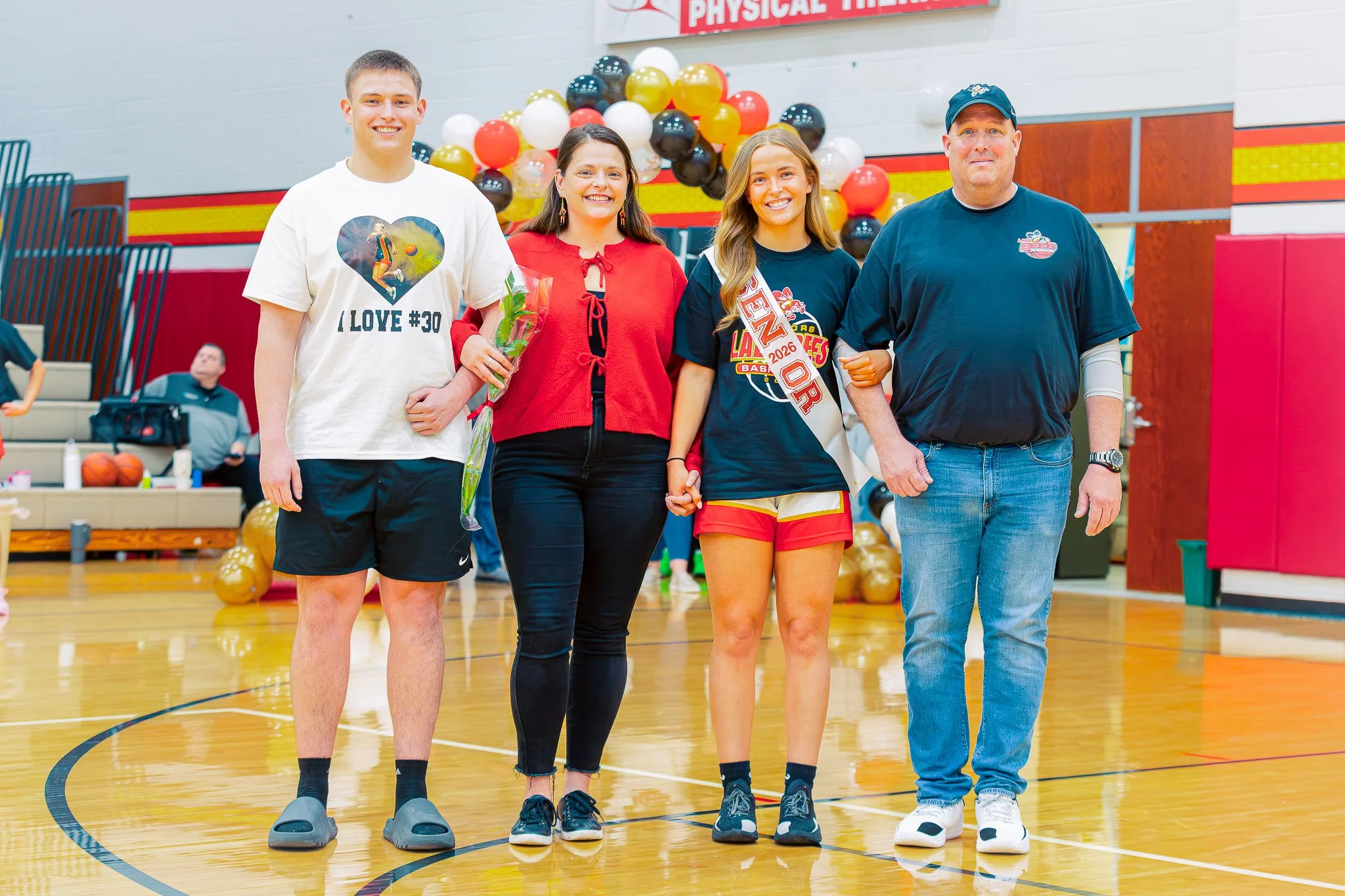 Four people standing in a gymnasium with balloons in the background. Basketball Senior Night.