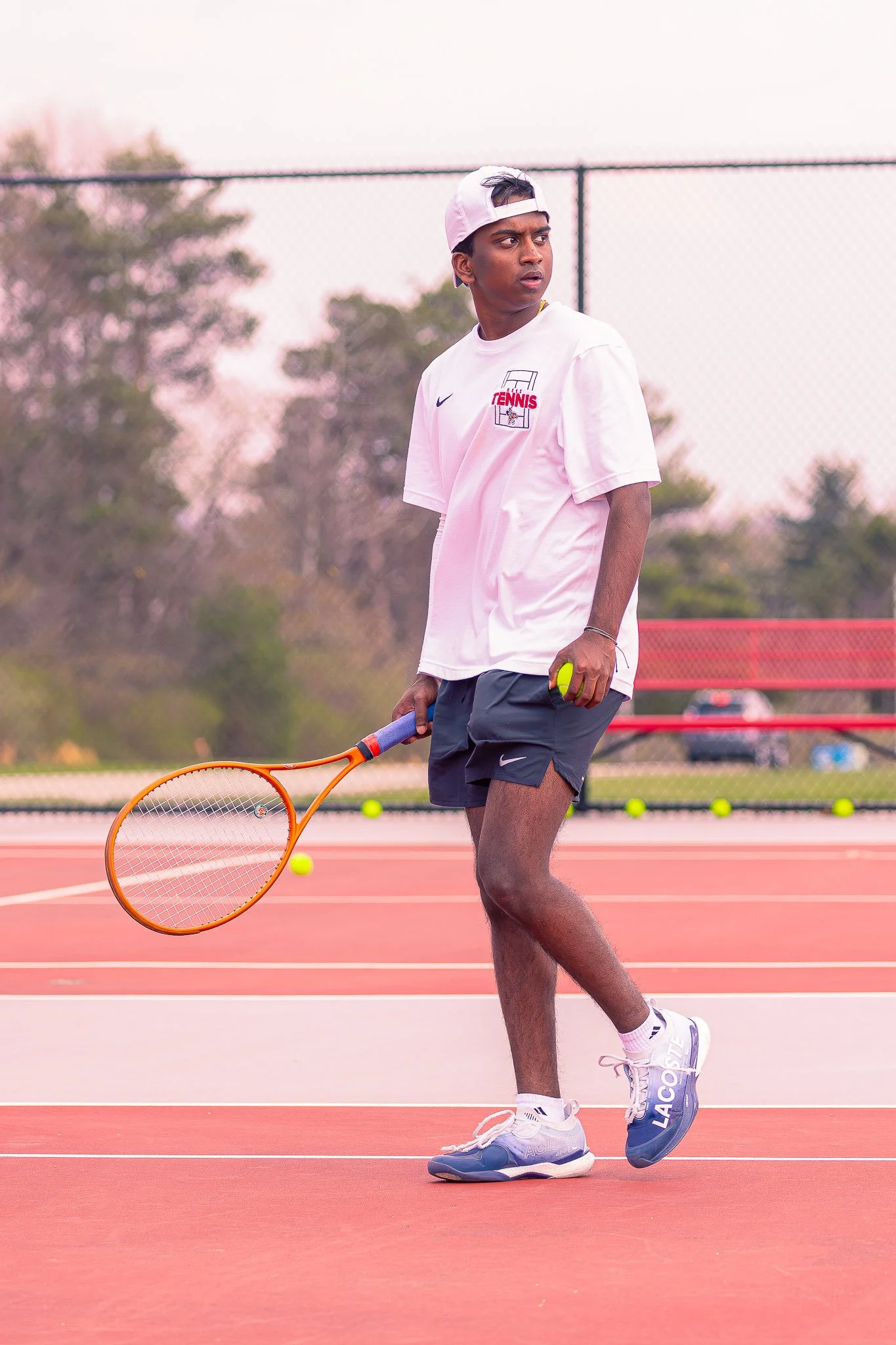 A young male tennis player standing on a tennis court, holding a tennis racket, wearing a white cap, white t-shirt with a tennis logo, dark shorts, and blue tennis shoes, with multiple tennis balls on the court around him.