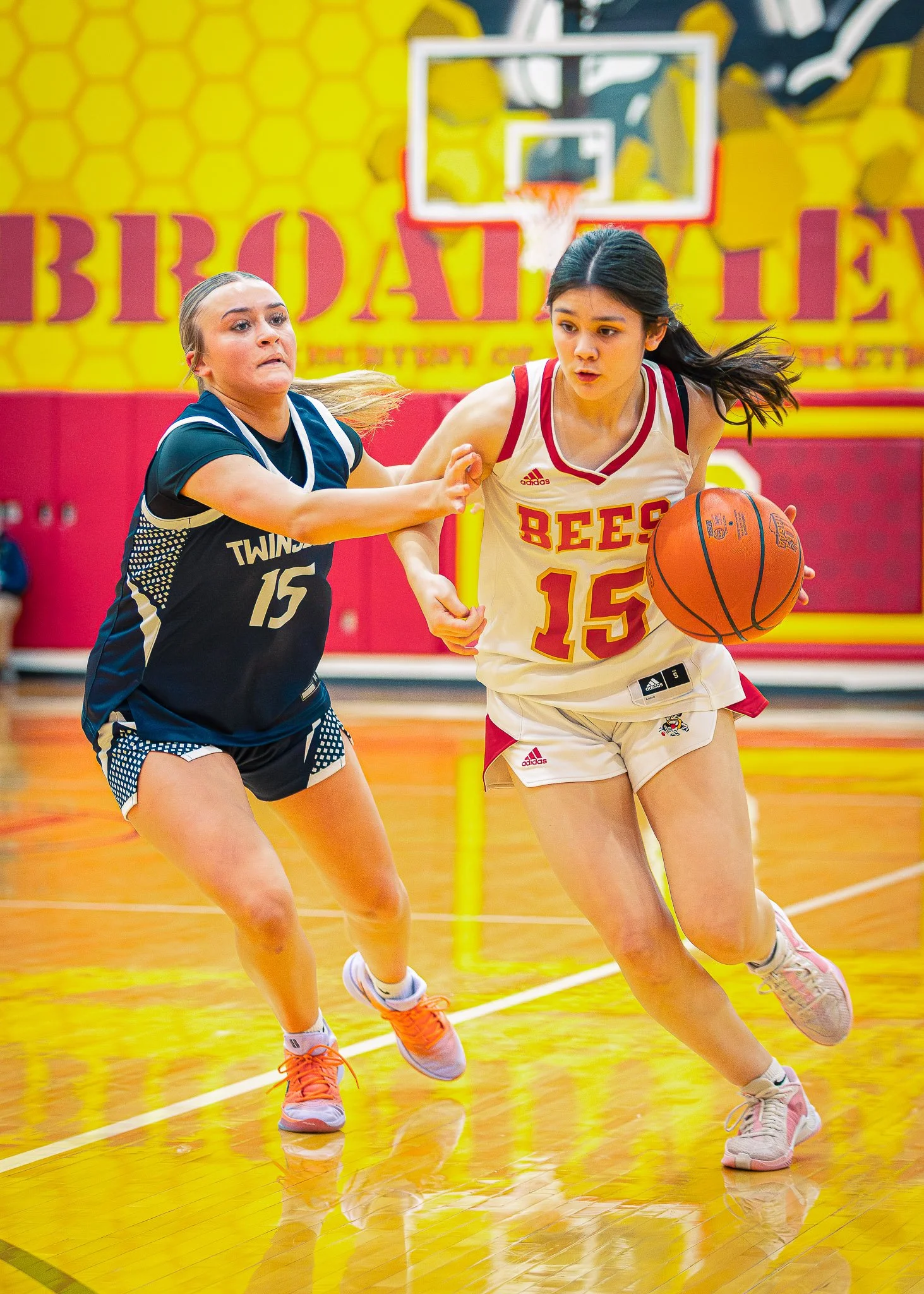 Two female basketball players with athletic builds compete for possession of the basketball during a game in a gymnasium. The player on the right wears a white and red uniform, dribbling the ball, while the player on the left wears a dark blue and wh